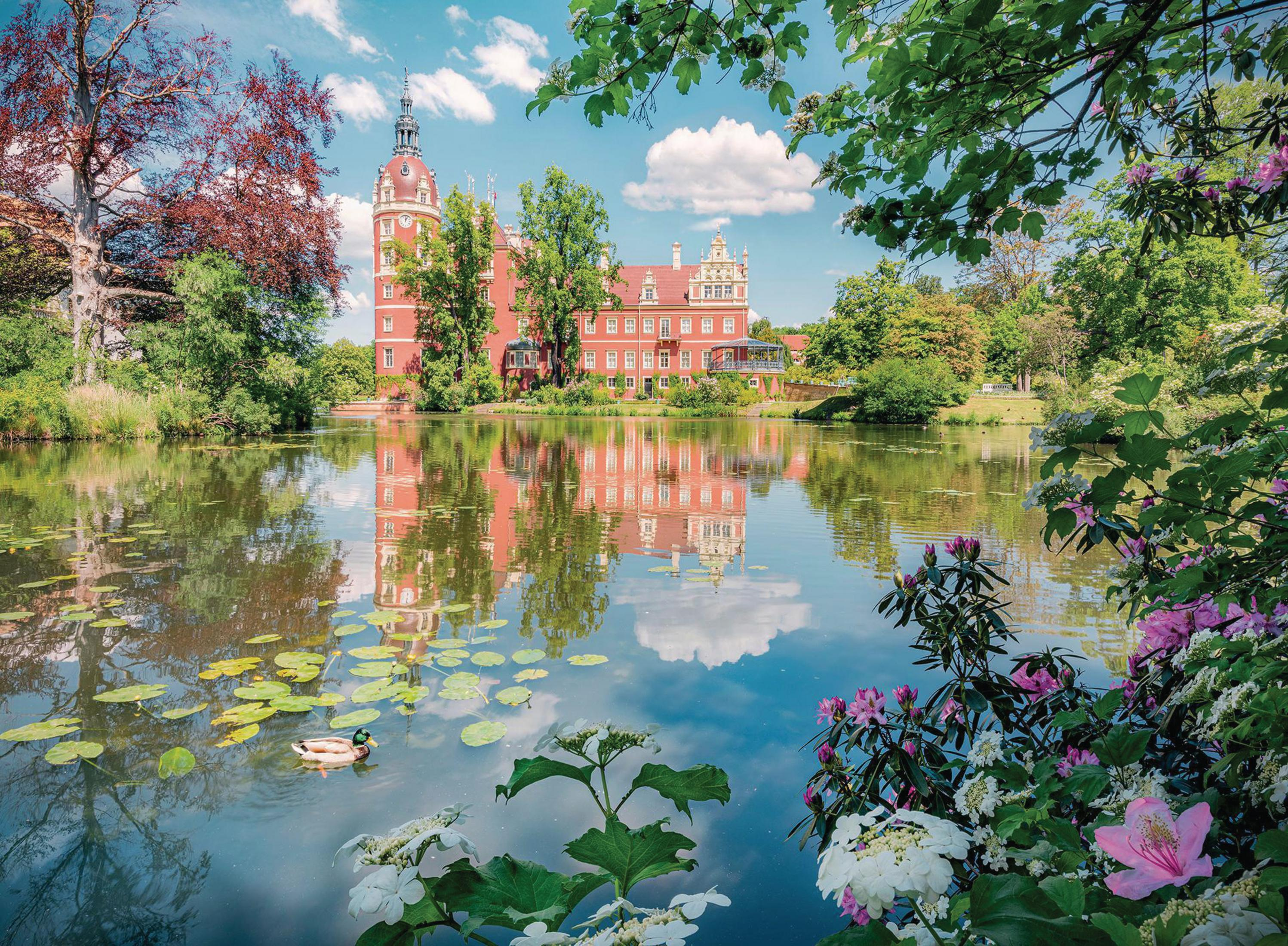 Ein rotes Schloss an einem See, das sich im Wasser spiegelt. Grüne Bäume umgeben die Szene unter blauem Himmel mit Wolken.