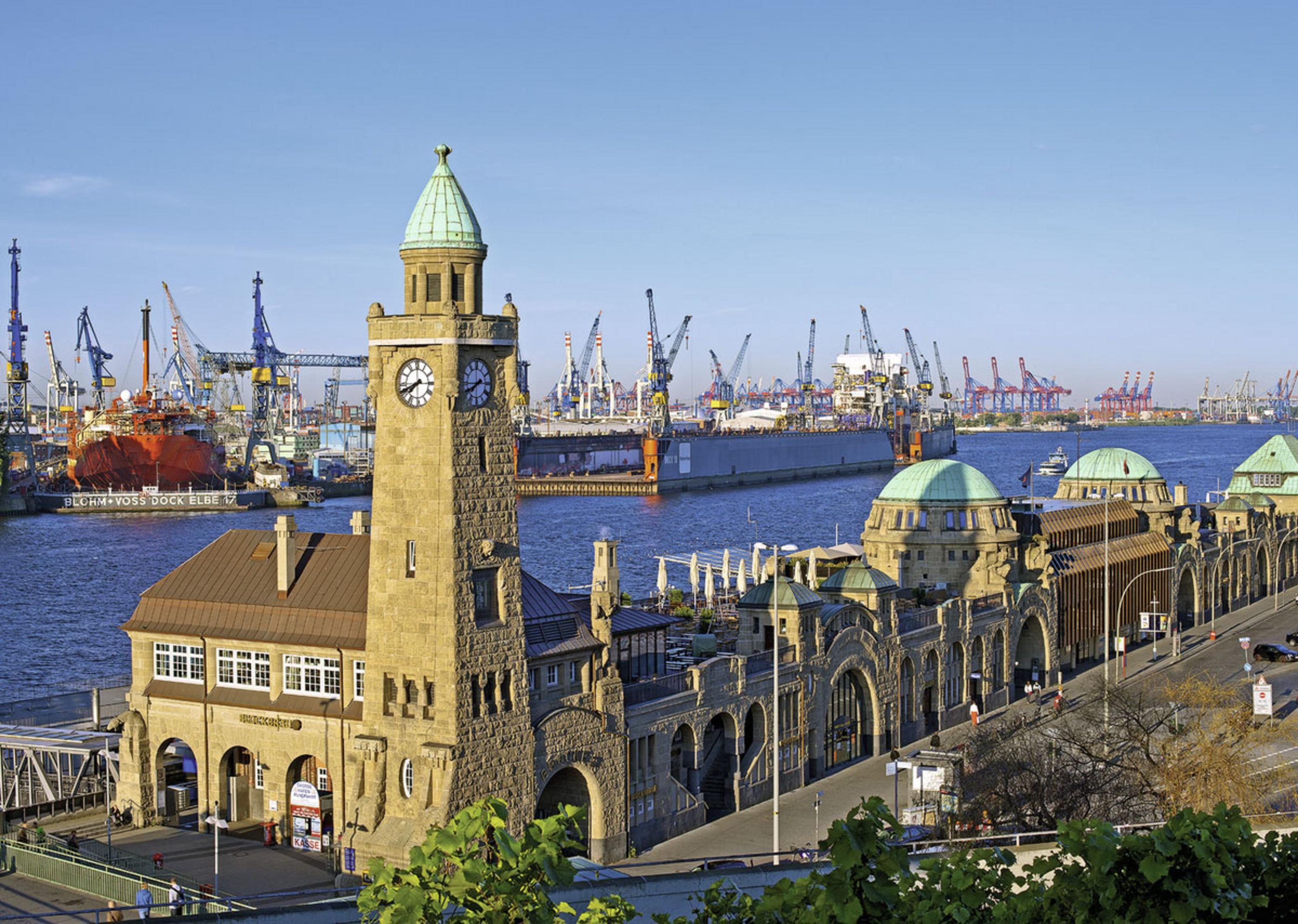 Ein Uhrturm am Hafen, Kräne, Schiffe und Gebäude füllen den Hintergrund unter blauem Himmel.