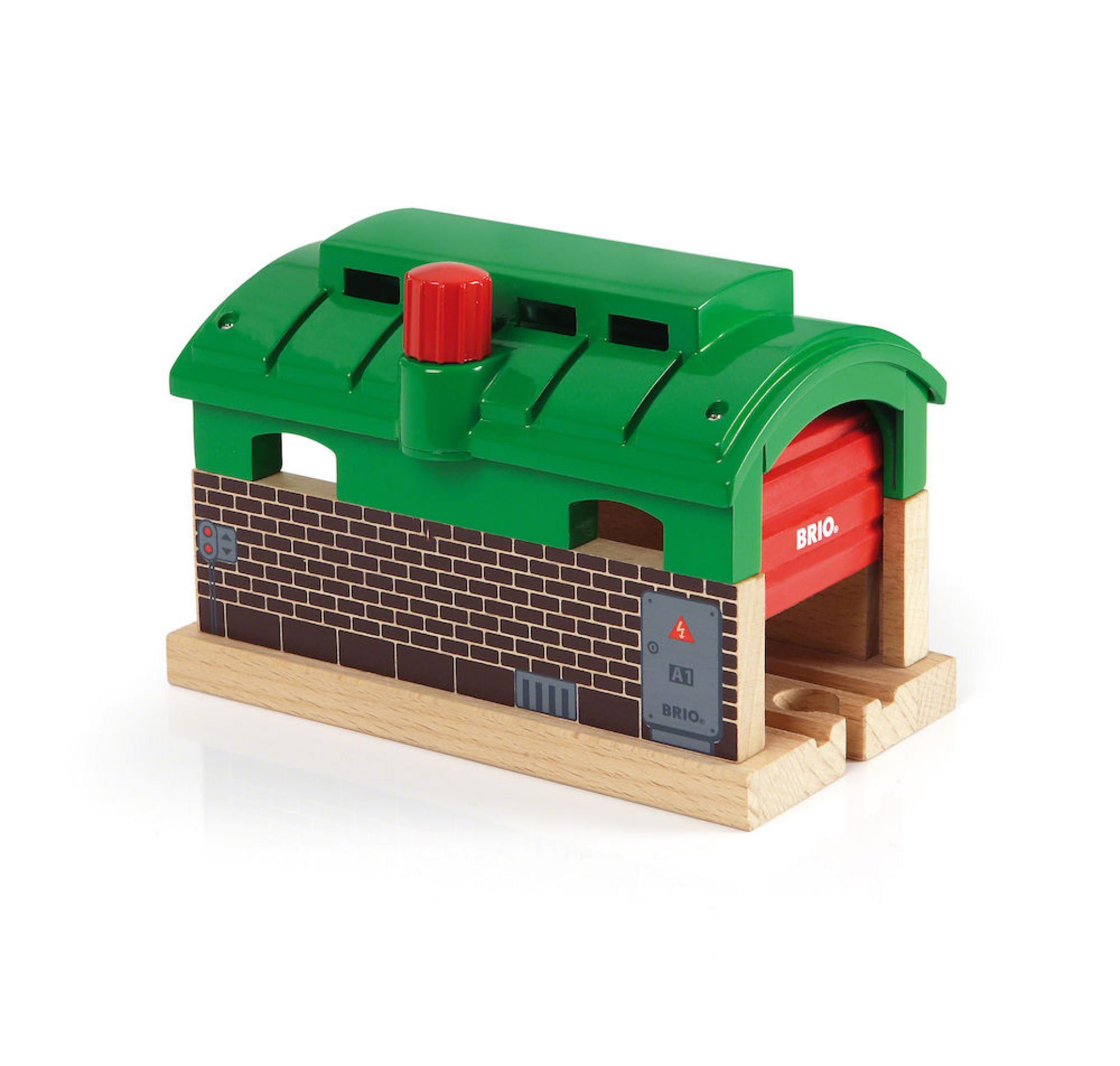 Un cobertizo de tren de juguete verde con una puerta roja está sobre vías de madera. El cobertizo tiene una chimenea roja.