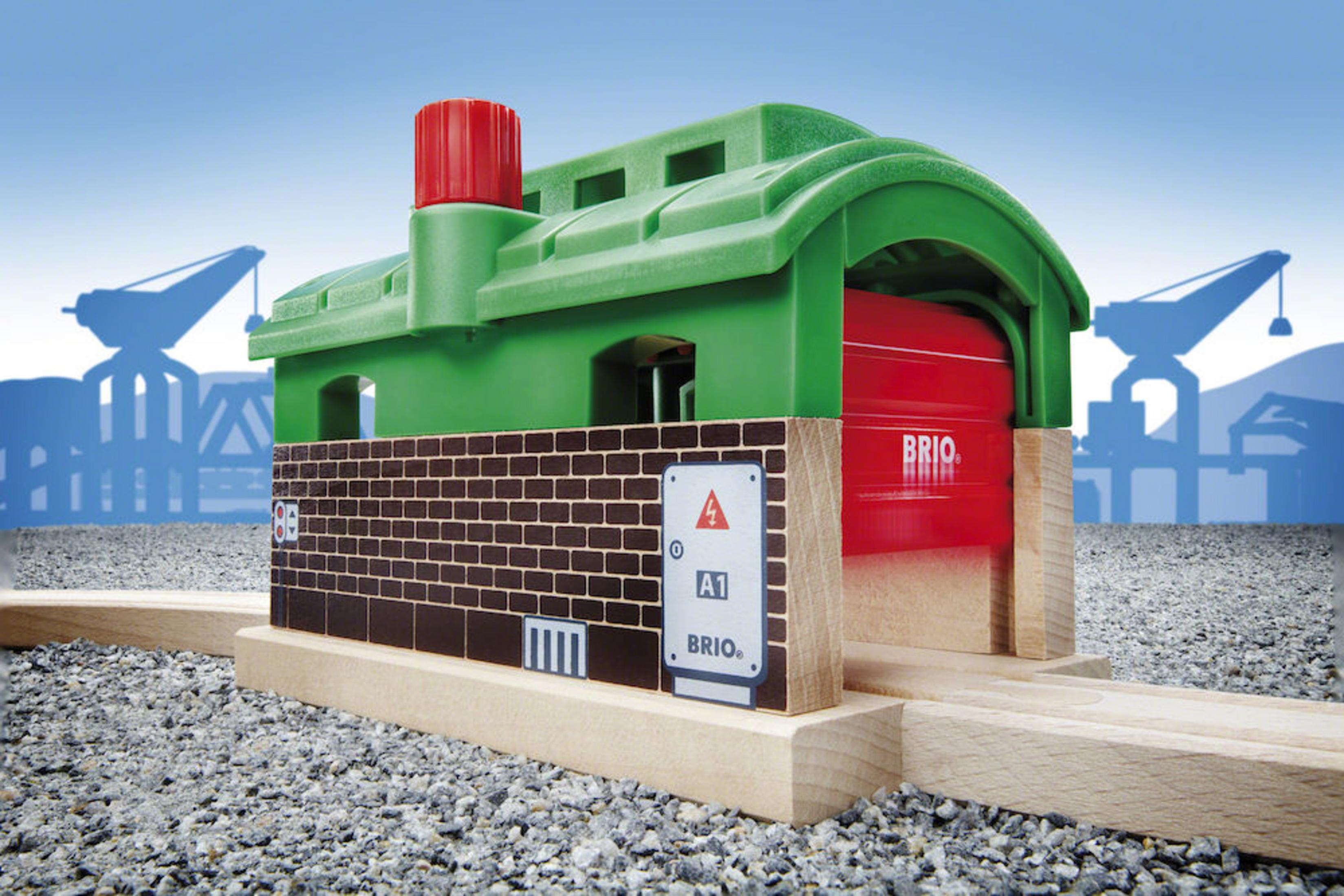 Un cobertizo de tren de juguete con techo verde y puerta roja. Está sobre un suelo gris.