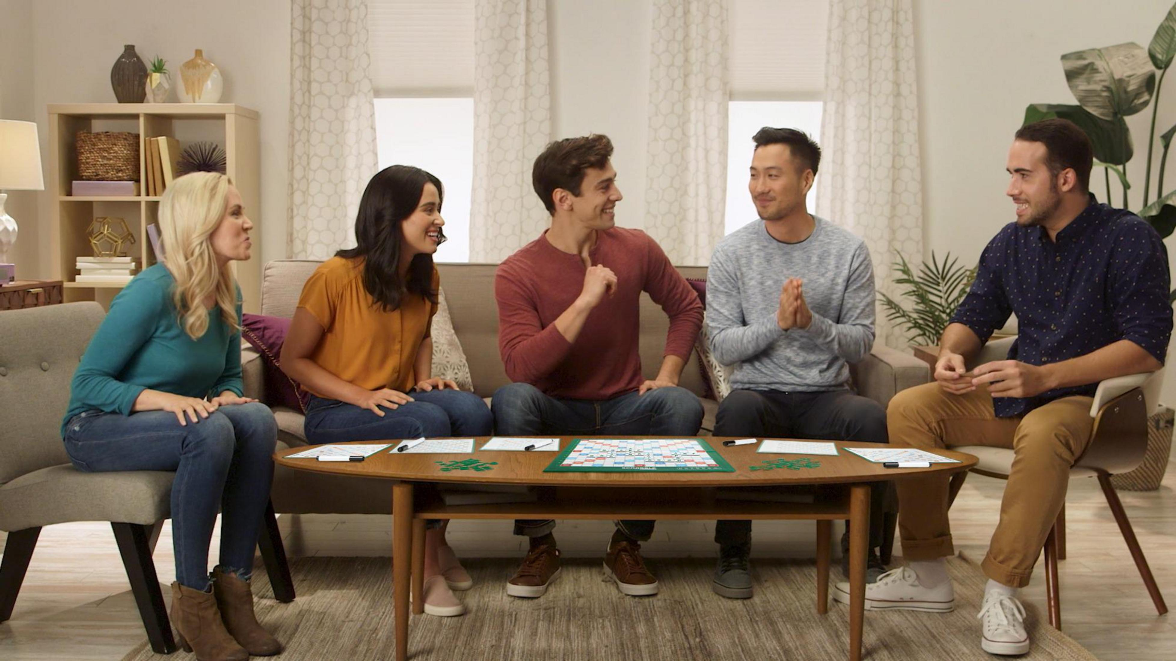 Pięć osób siedzi przy stole, grając w grę. Interagują i patrzą na siebie.