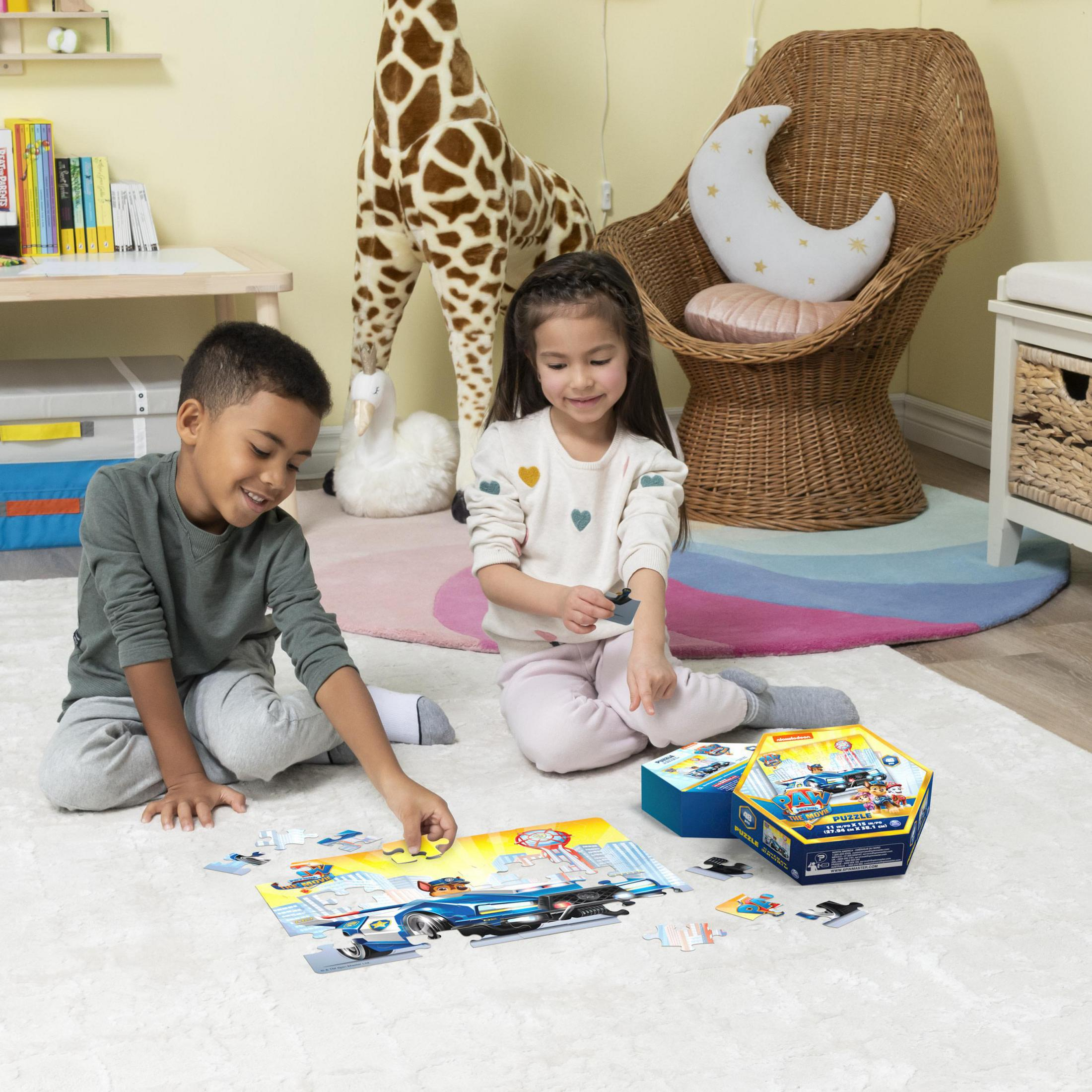 Zwei Kinder spielen ein Puzzle auf einem Teppich. Ein Giraffe und ein Stuhl sind im Hintergrund.