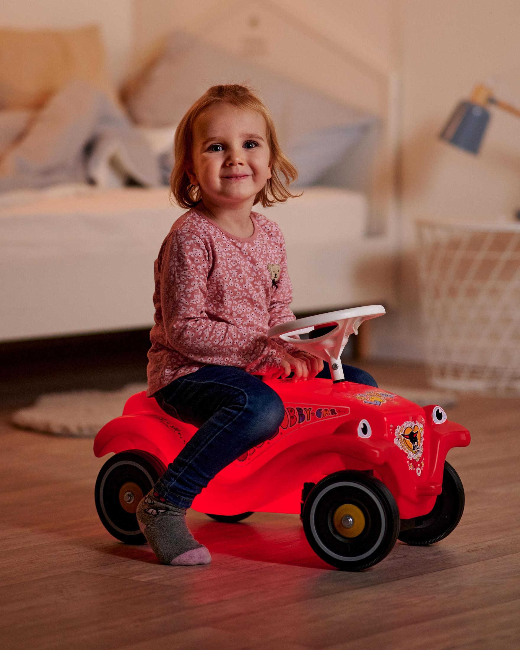 Una niña pequeña sentada en un coche de juguete rojo sonriendo, iluminada por una luz cálida.
