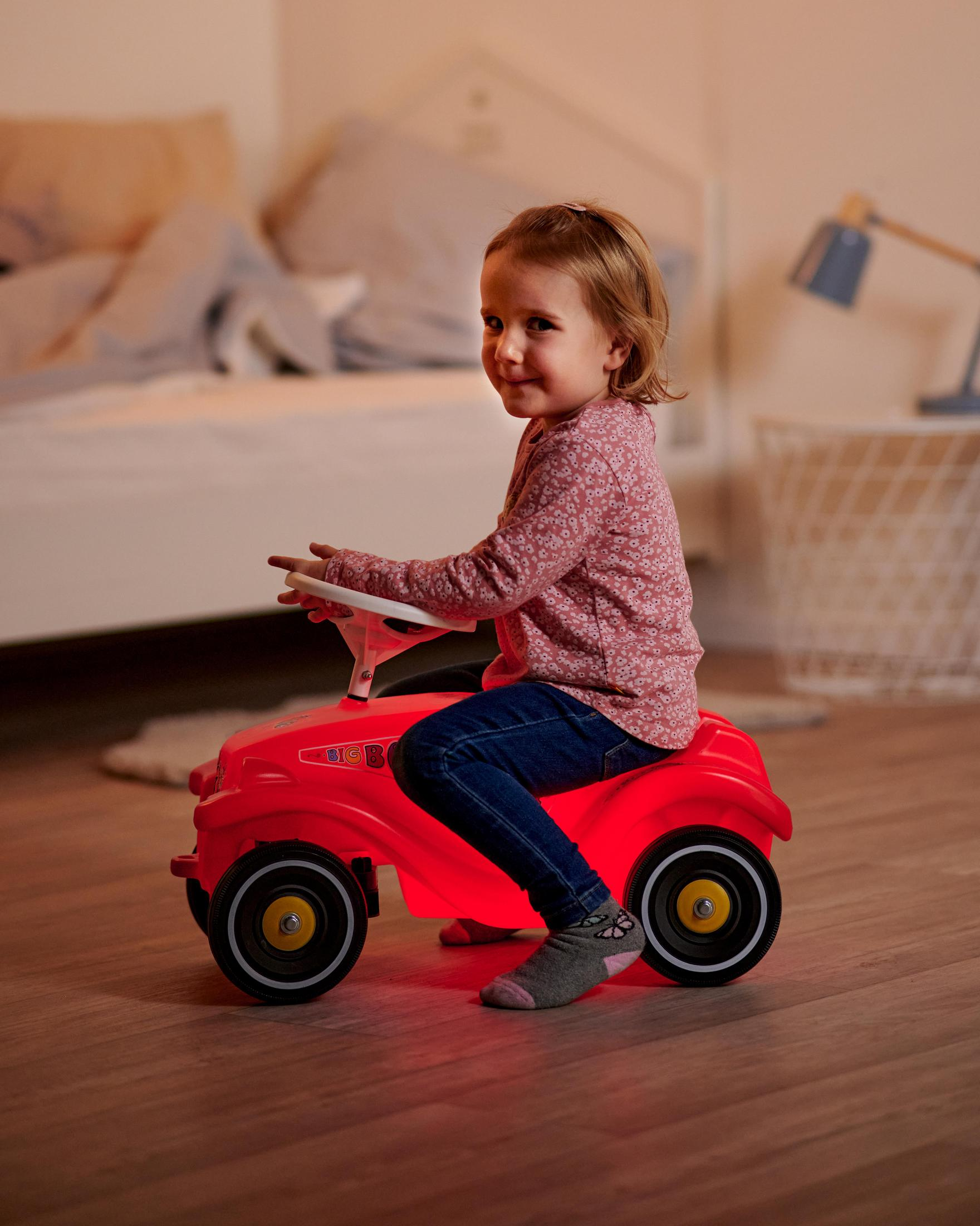 Una niña pequeña en un coche de juguete rojo. Está en una habitación con una cama y una lámpara.