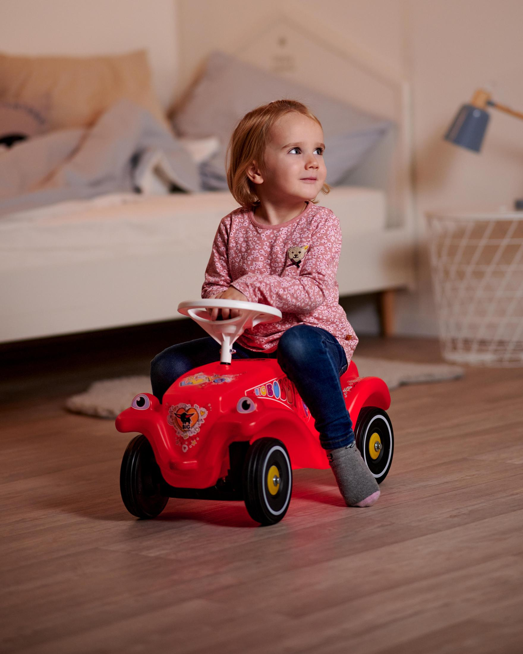 Una niña pequeña monta un coche de juguete rojo en una habitación. Mira hacia arriba y sonríe.