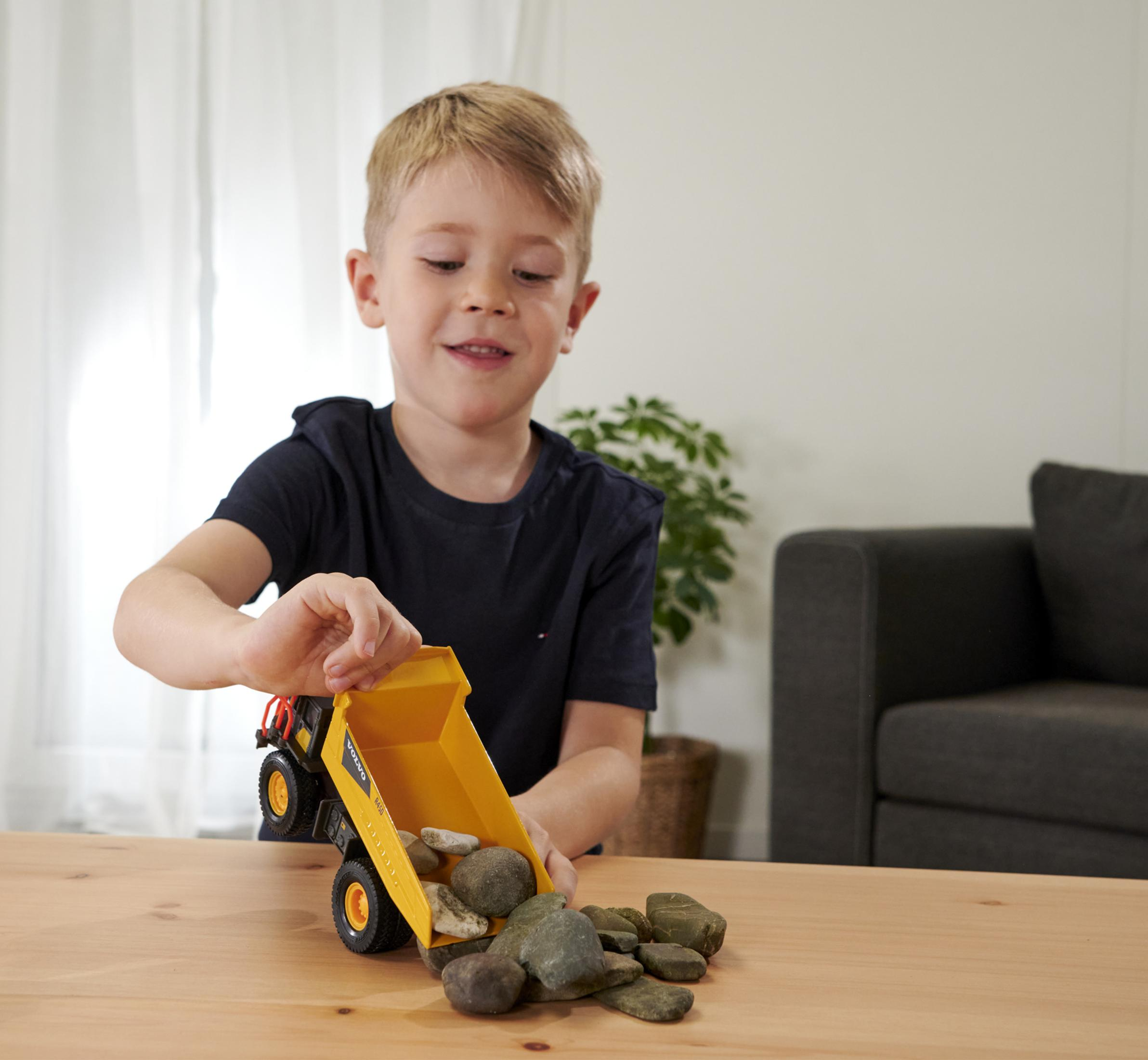 Mały chłopiec bawi się żółtą zabawką wywrotką, wysypując kamienie na drewniany stół. Z tyłu roślina.