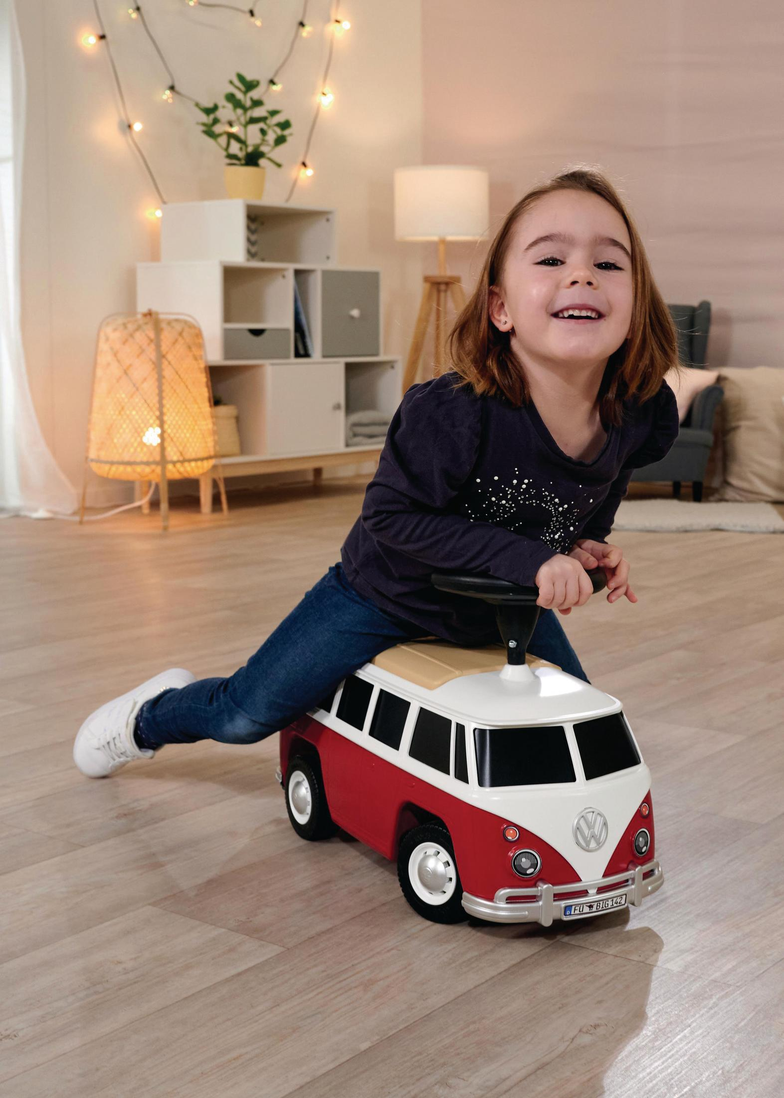 Ein junges Mädchen fährt auf einem rot-weißen Spielzeug-VW-Bus. Sie lächelt. Der Boden ist aus Holz.