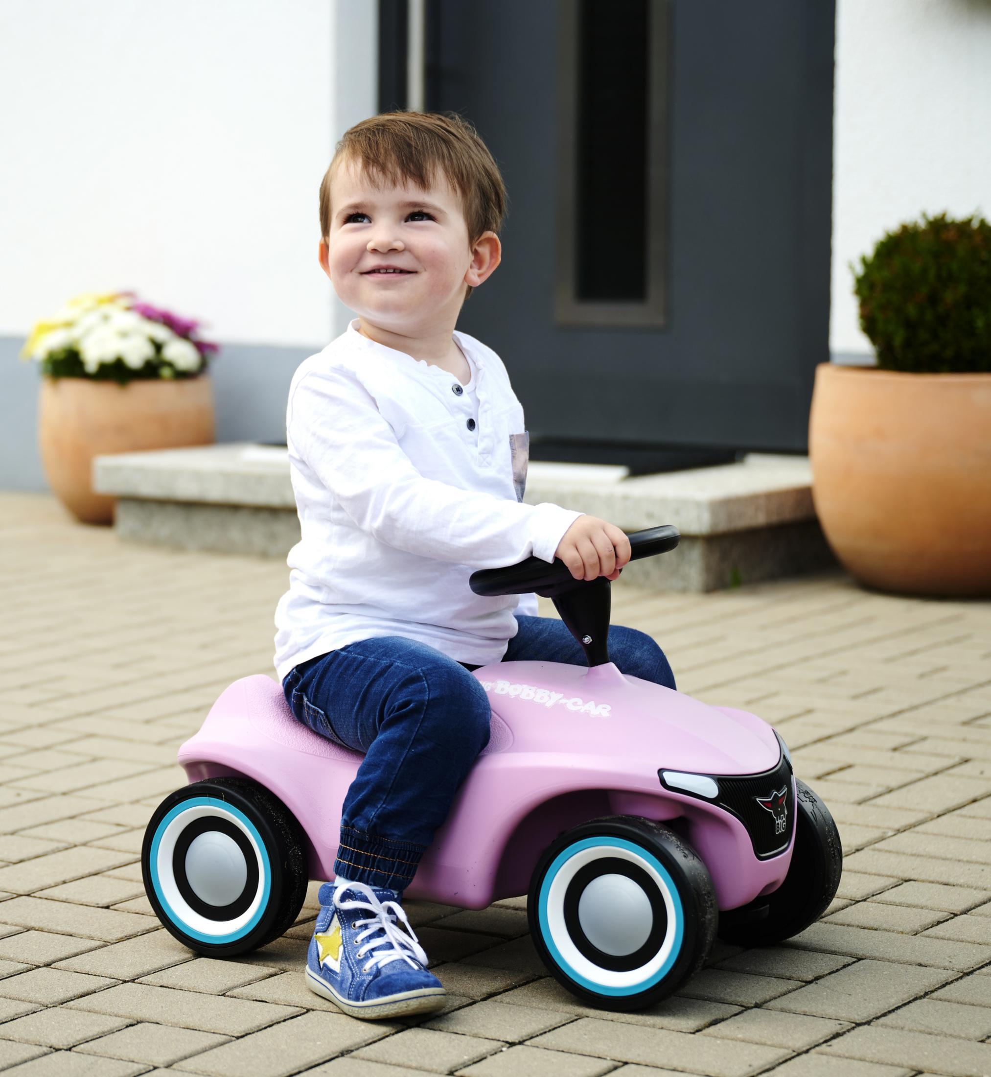 Ein Junge in weißem Hemd und Jeans sitzt auf einem rosa Bobby-Car und lächelt. Hintergrund: Haus, Pflanzen.