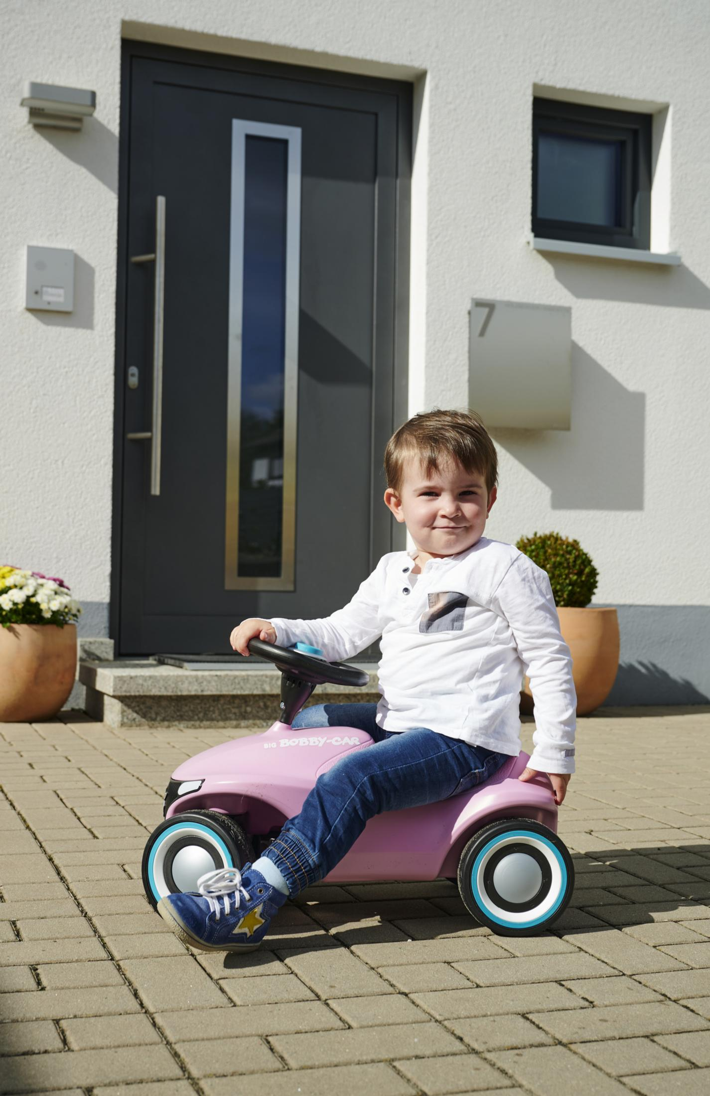 Ein kleiner Junge sitzt in einem rosa Spielzeugauto vor einem weißen Haus.