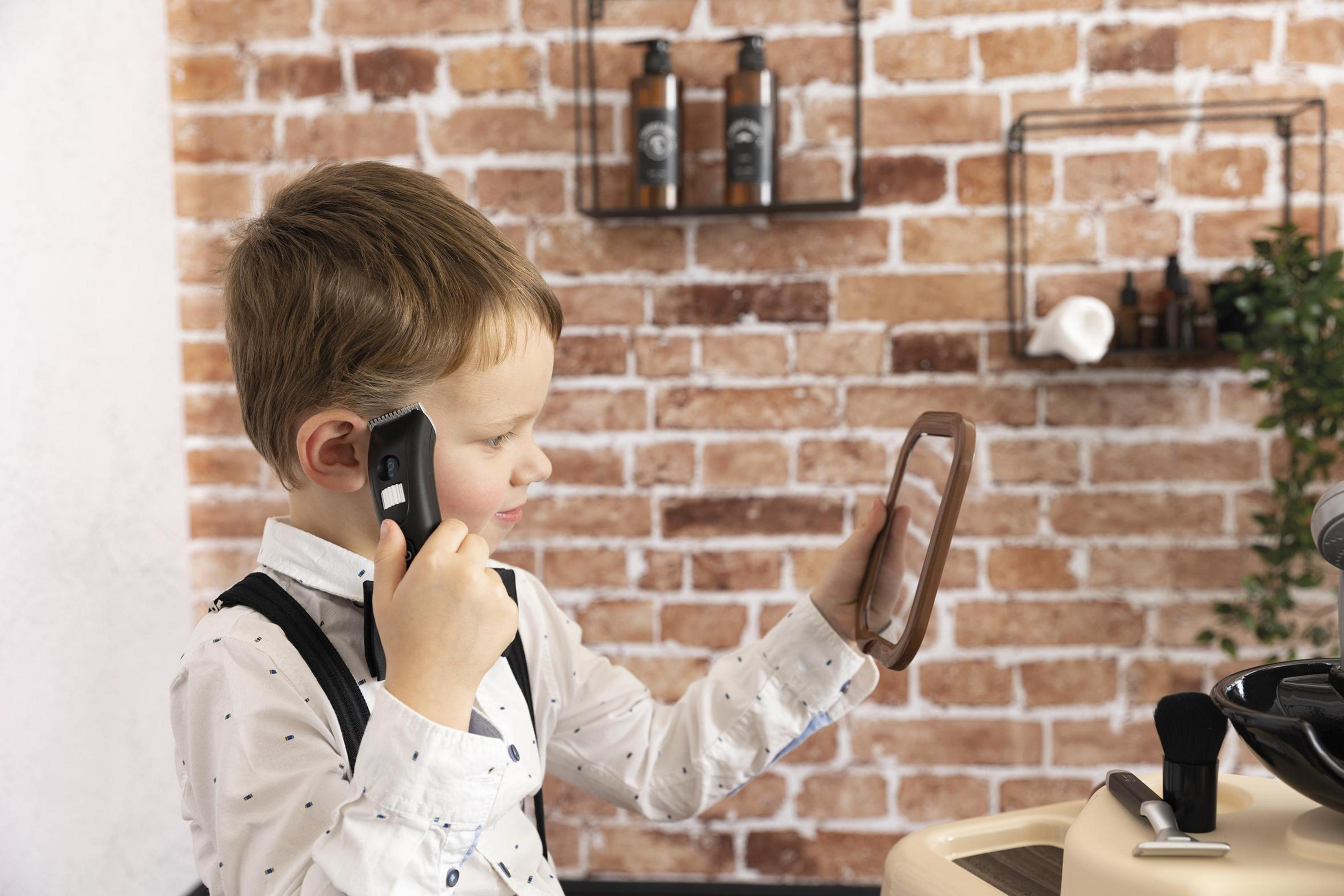 Ein kleiner Junge schneidet sich mit einem Haarschneider die Haare und schaut in einen Spiegel. Eine Backsteinmauer ist der Hintergrund.