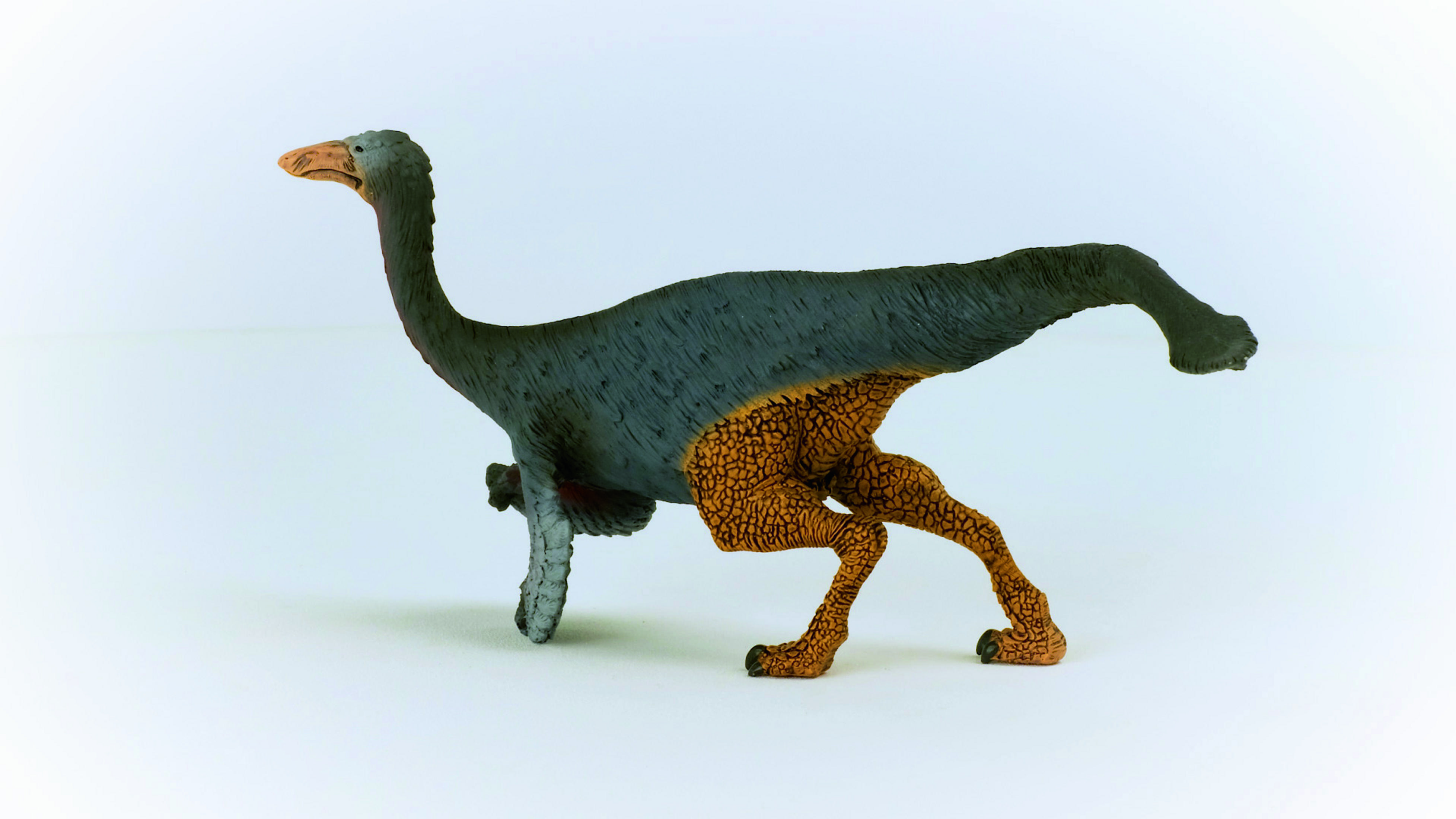 Zabawka dinozaura z szarym ciałem, pomarańczowymi nogami i brązowym dziobem, na białej powierzchni.