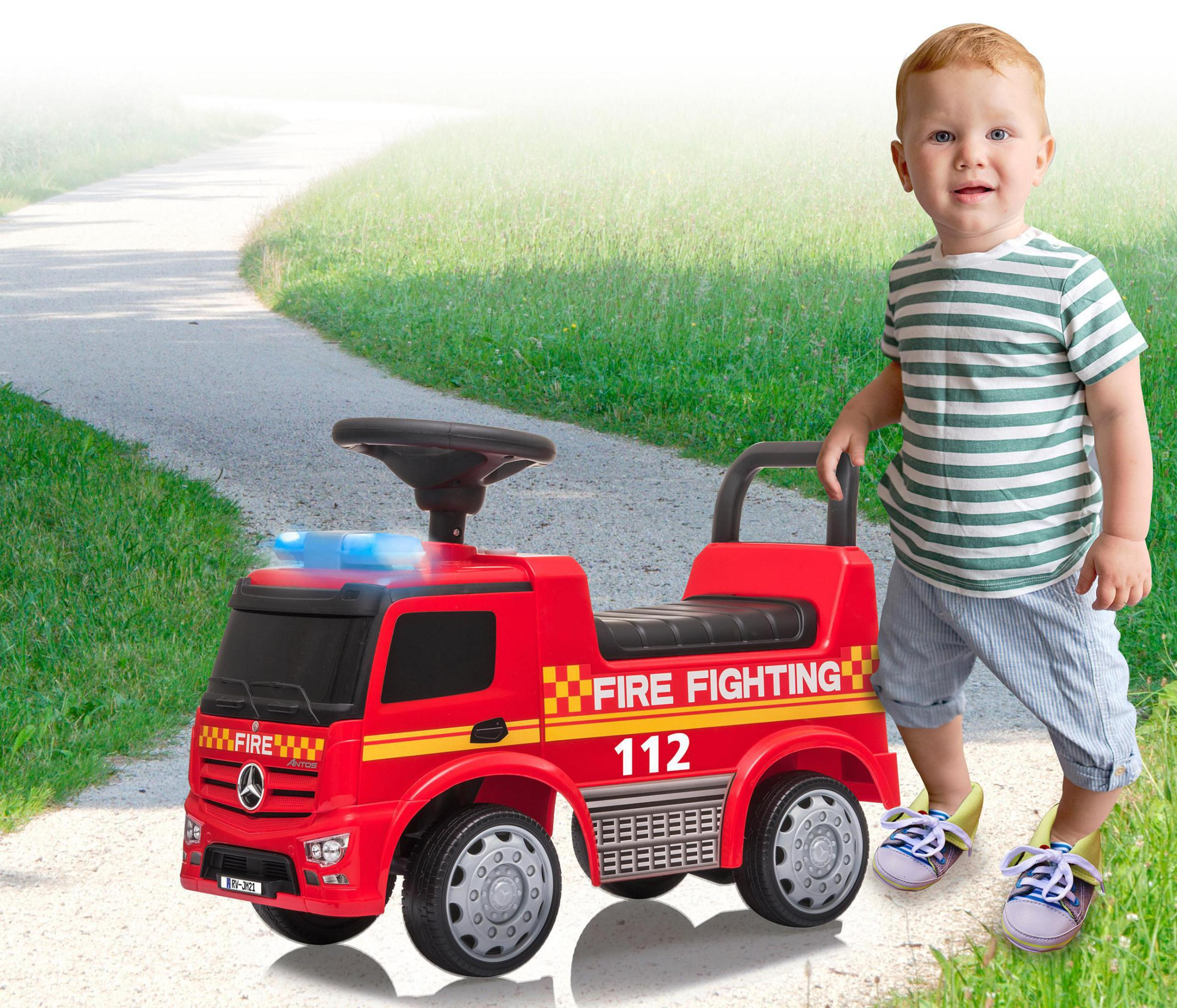 Ein kleiner Junge steht neben einem roten Feuerwehrauto-Spielzeug. Er lächelt und trägt ein gestreiftes Hemd.