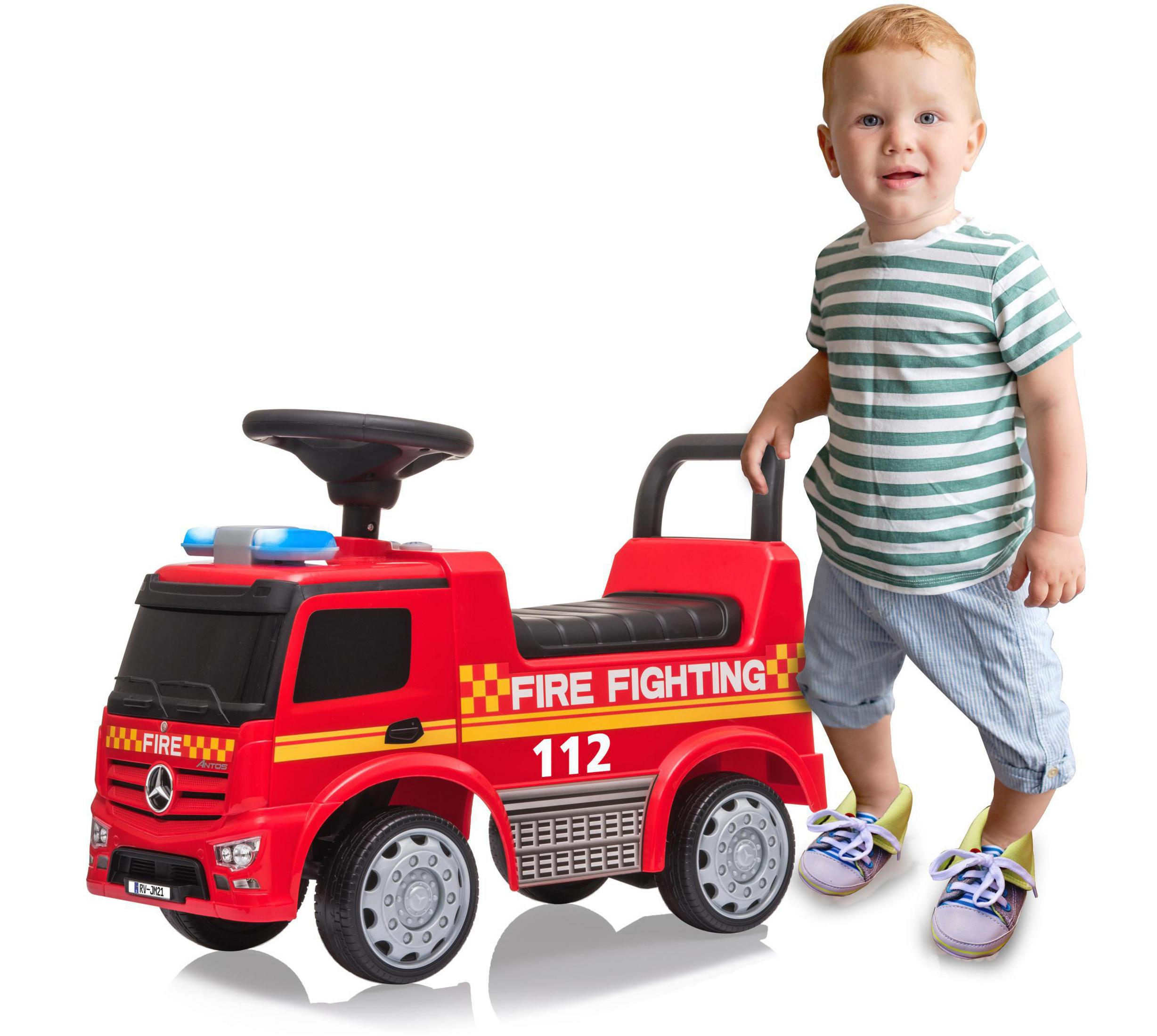 Ein Kleinkind steht neben einem roten Spielzeug-Feuerwehrauto. Auf dem LKW steht 'FIRE FIGHTING 112'.