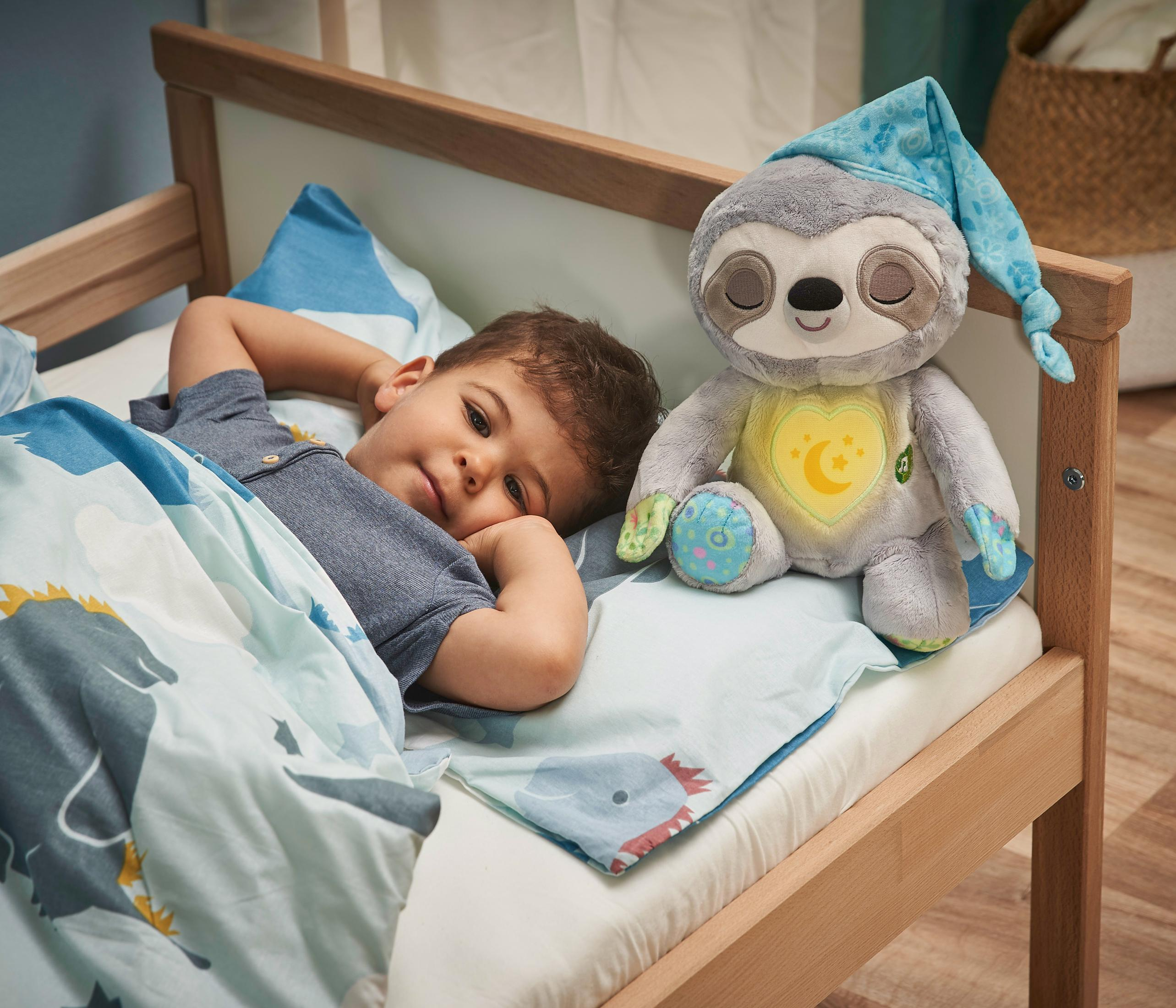 Ein kleiner Junge liegt im Bett, neben einem Stofffaultier mit Nachtmütze. Der Raum ist hell und luftig.