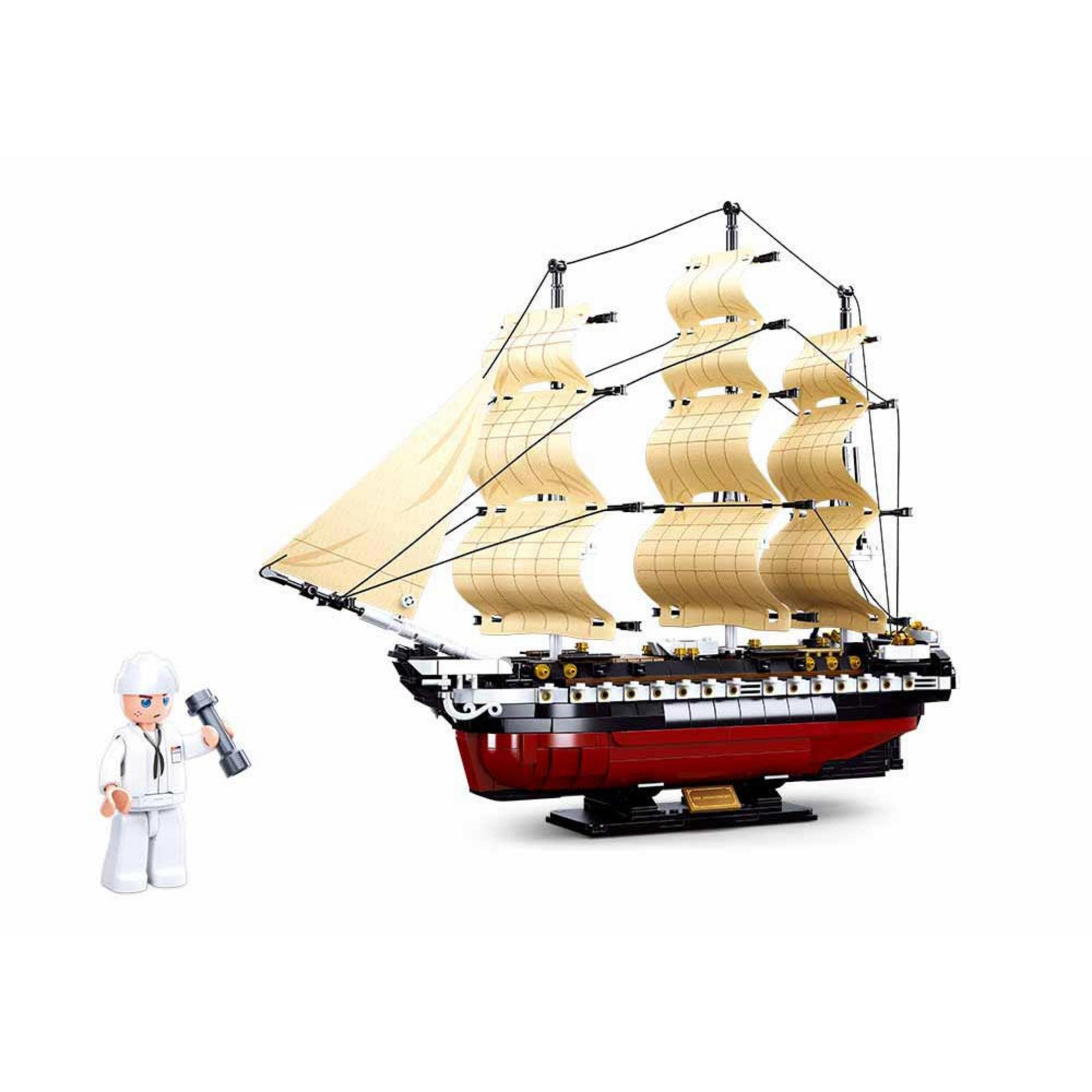 Modellschiff mit beigen Segeln und rotem Rumpf. Figur hält Taschenlampe, stehend.