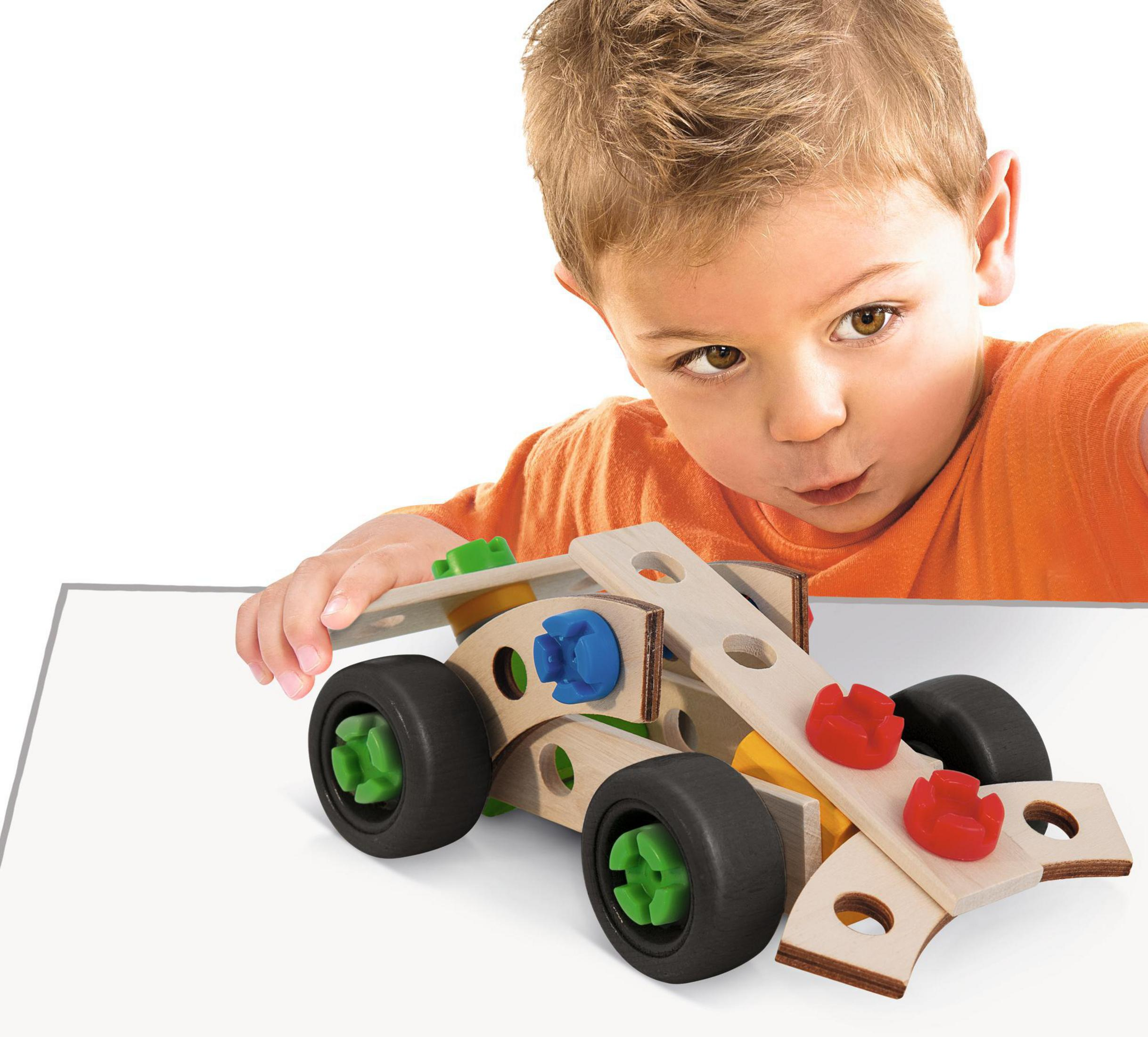 Junge betrachtet ein Holzspielzeugauto. Das Auto hat schwarze Räder und rote und grüne Teile.