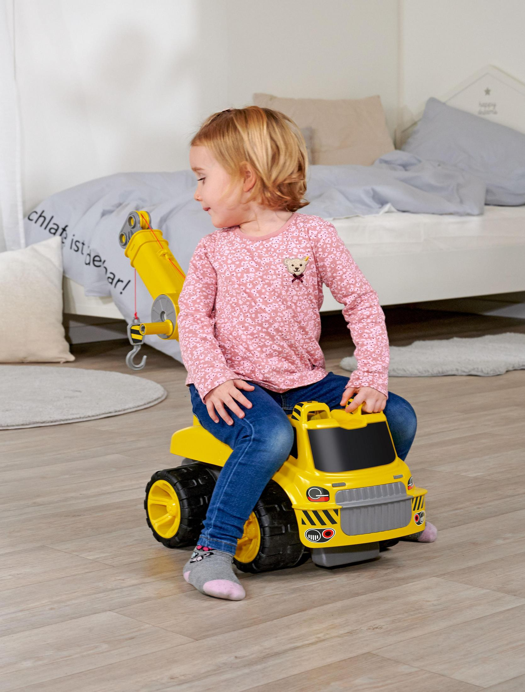 Mała dziewczynka siedzi na żółtej zabawce ciężarówki. Patrzy w lewo.