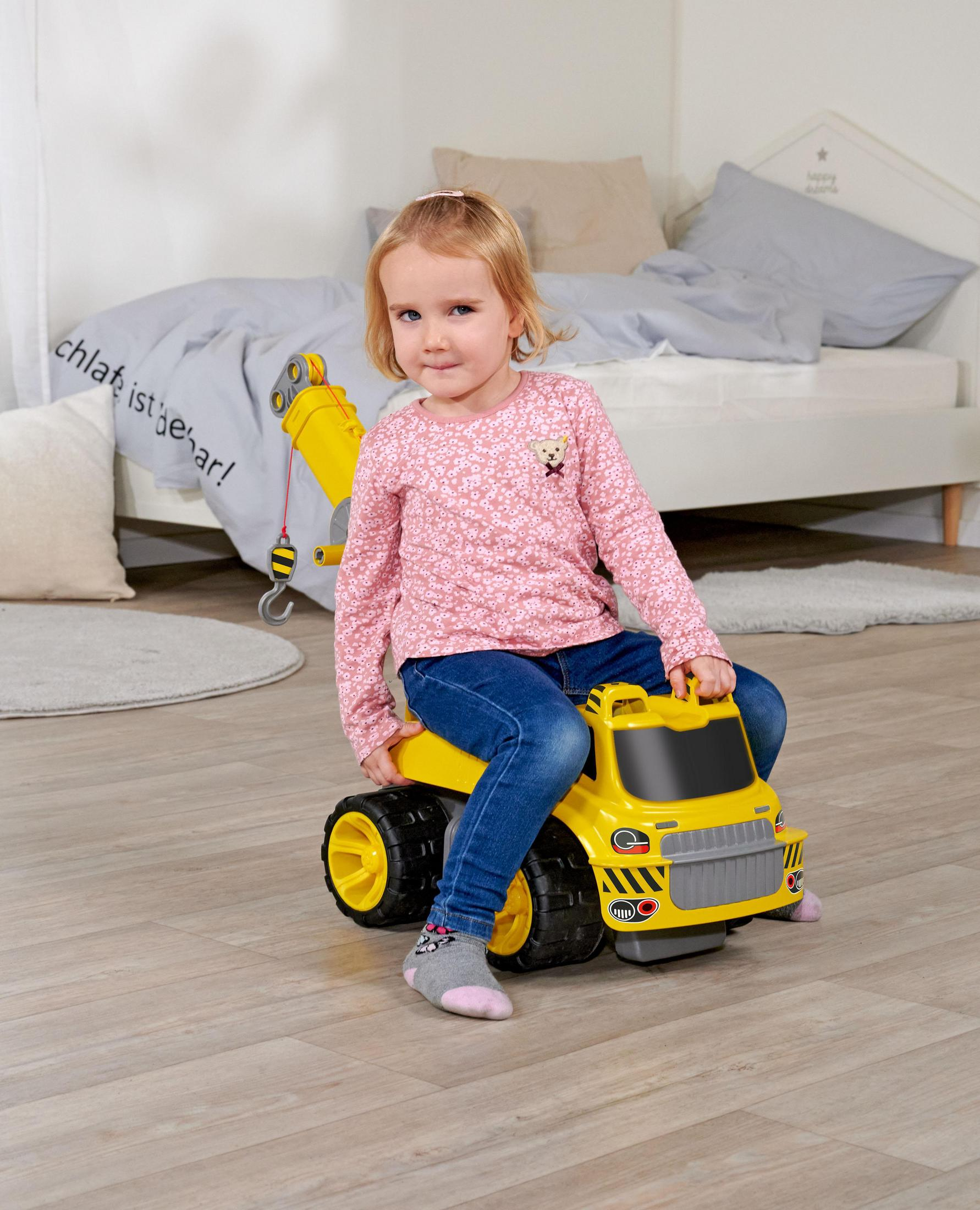Mała dziewczynka w różowej bluzce siedzi na żółtej zabawce-ciężarówce. Jasna drewniana podłoga, białe tło ściany.