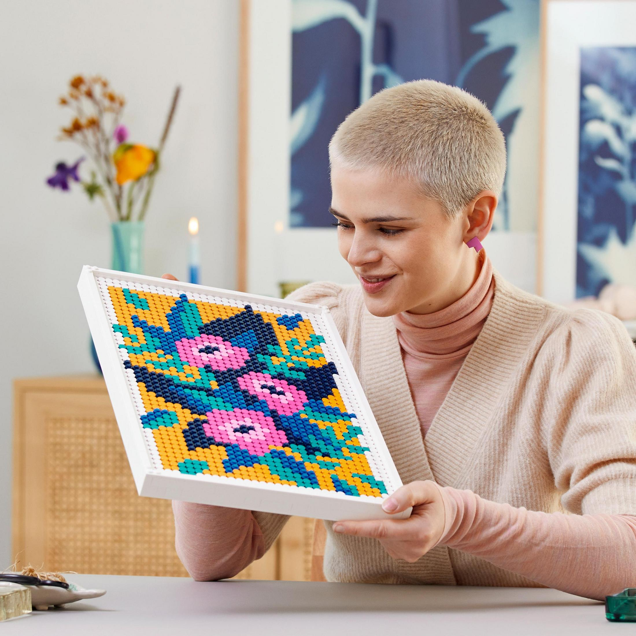 Mujer sosteniendo un cuadro de arte LEGO enmarcado, patrón de flores coloridas. Sonríe.