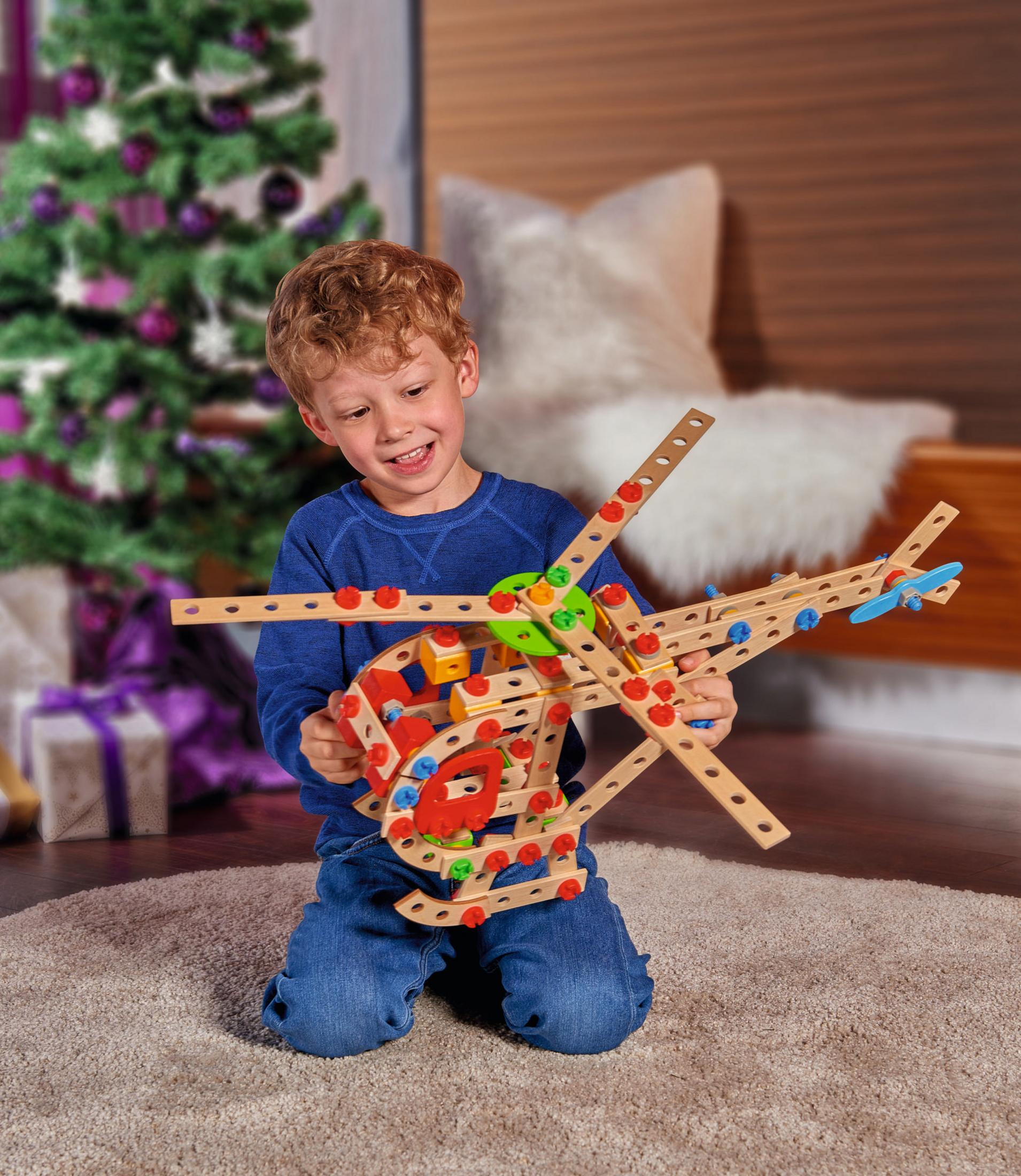 Ein Junge kniet und hält einen selbstgebauten Holzspielzeughubschrauber, lächelnd.