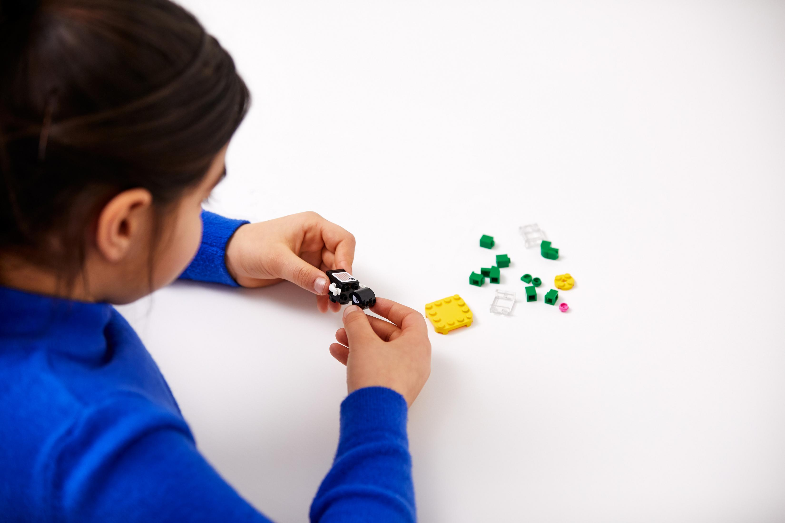 Una persona che assembla pezzi Lego su una superficie bianca. Ci sono pezzi sparsi, una base gialla e un pezzo nero.