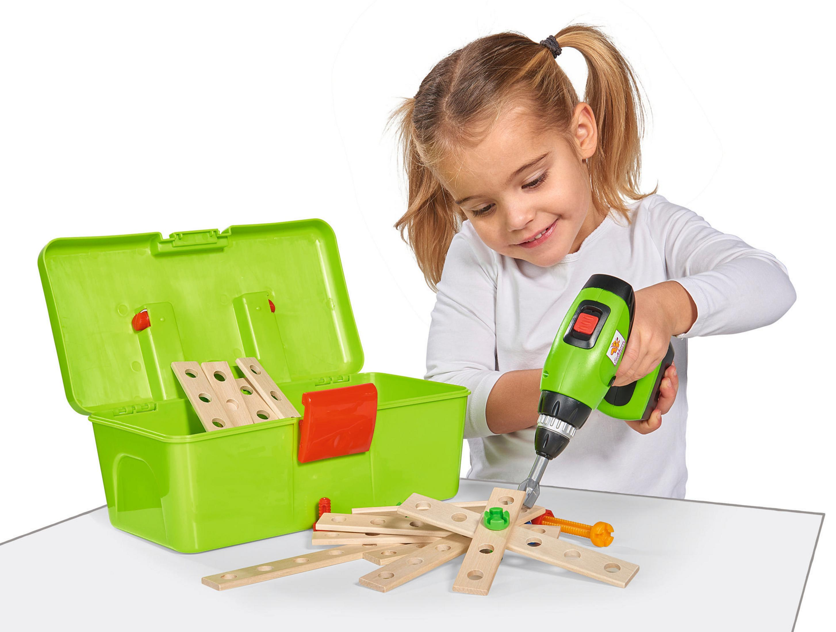 Ein junges Mädchen lächelt, während sie einen grünen Spielzeugbohrer auf Holzstücke verwendet, eine grüne Werkzeugkiste.