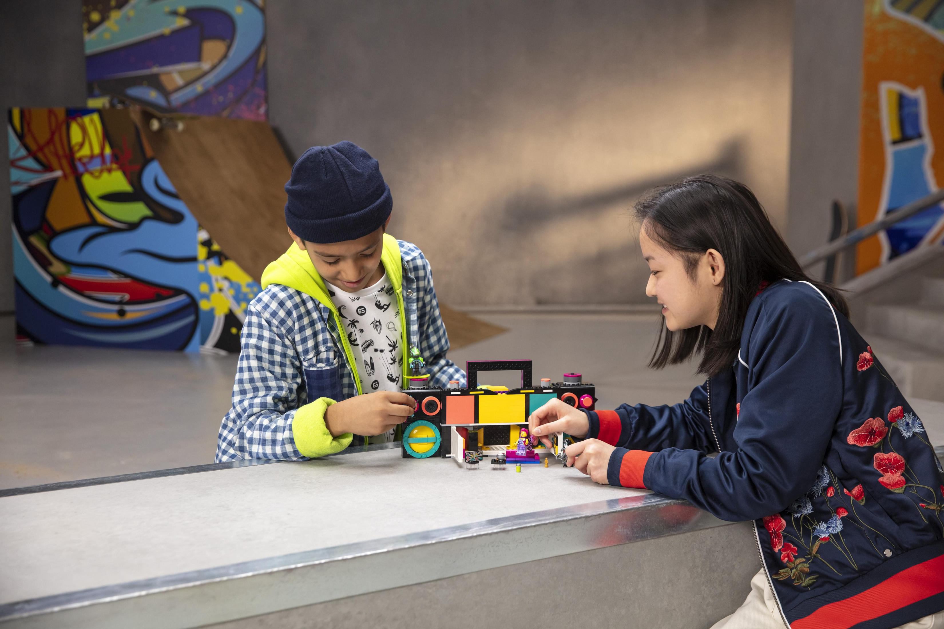 Dwoje dzieci buduje zestaw Lego w skateparku. Jedno trzyma element Lego, drugie patrzy.