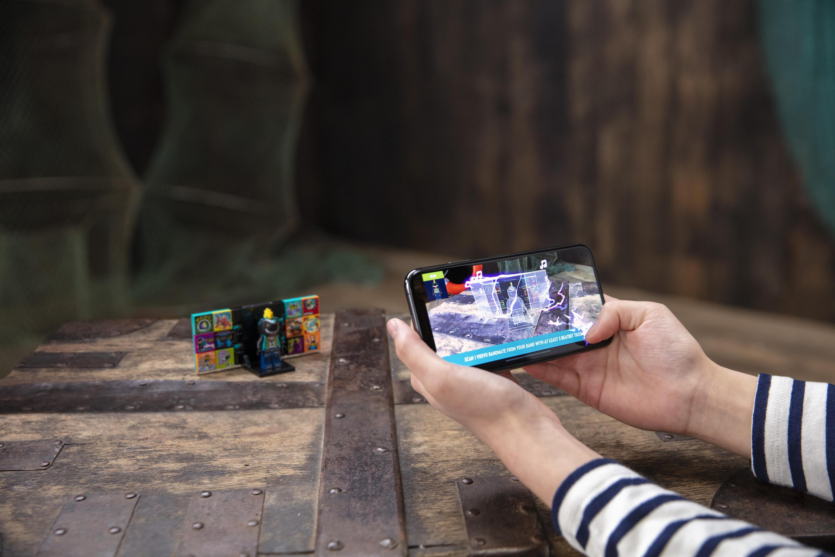 Osoba trzymająca telefon pokazujący scenę rozszerzonej rzeczywistości z klocków i figurki.