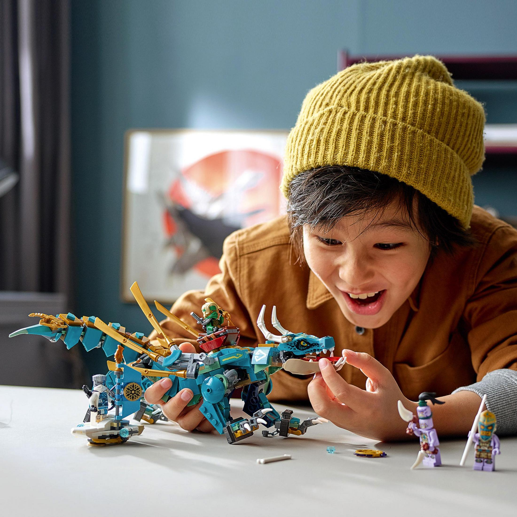 Bir çocuk Lego ejderha oyuncağıyla oynuyor. Sahnede Lego figürleri ve bir masa var.