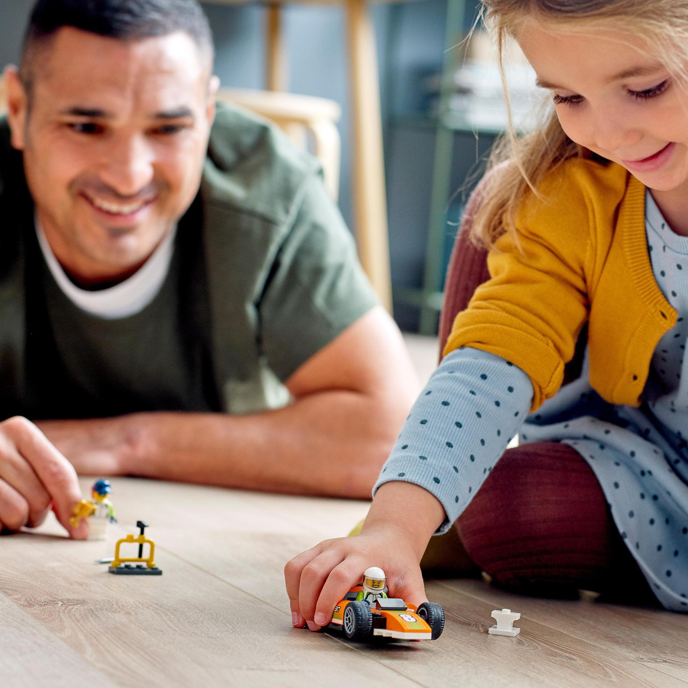 Een vader en kind spelen met Lego-auto's. Het kind houdt een oranje auto vast. De achtergrond is een houten vloer.