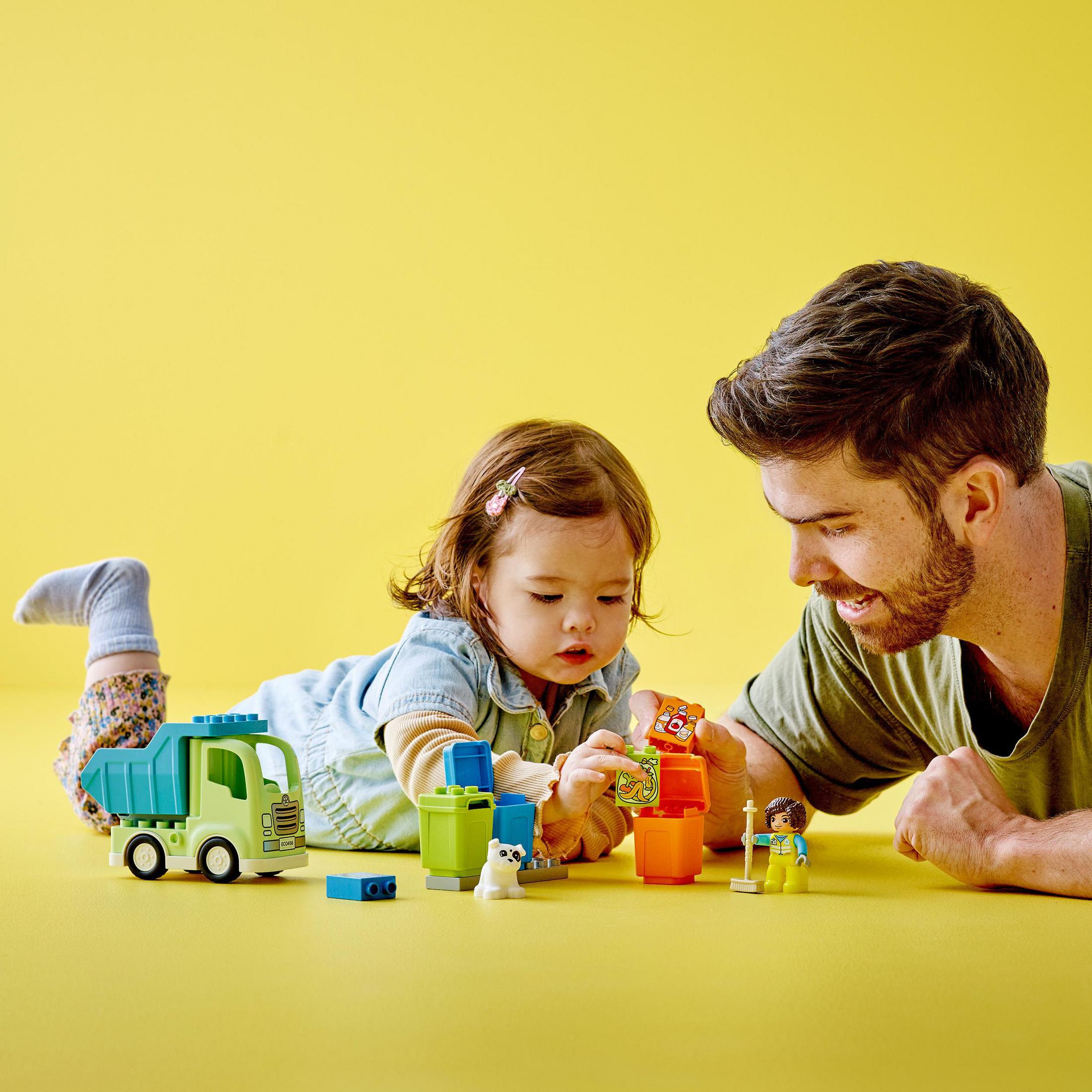 Ein Kleinkind und ein Mann spielen mit Spielzeugblöcken auf gelber Oberfläche, Spielzeug-LKW.