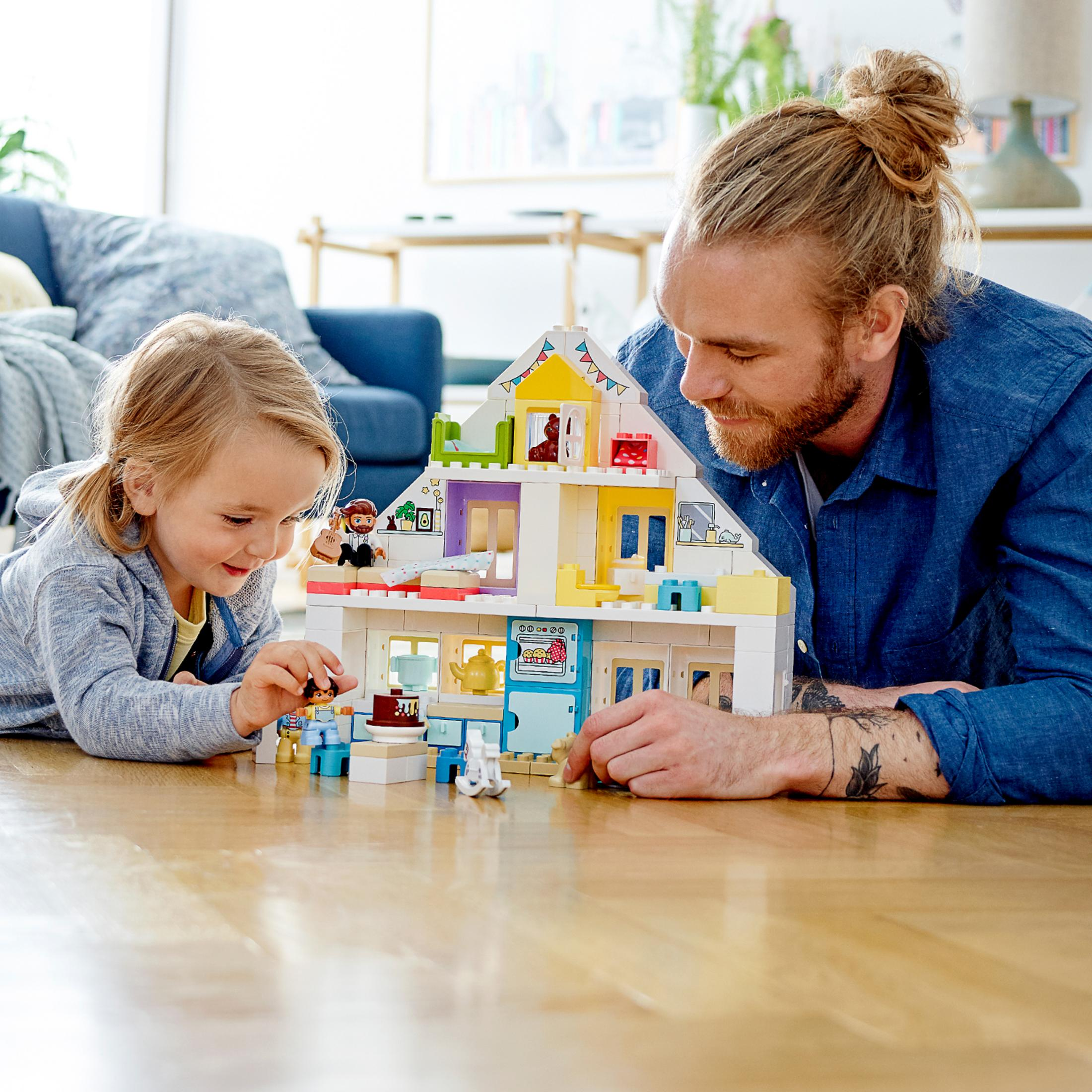 Un père et sa fille jouent avec une maison de poupée. La maison de poupée est blanche, jaune et bleue, sur un sol en bois.