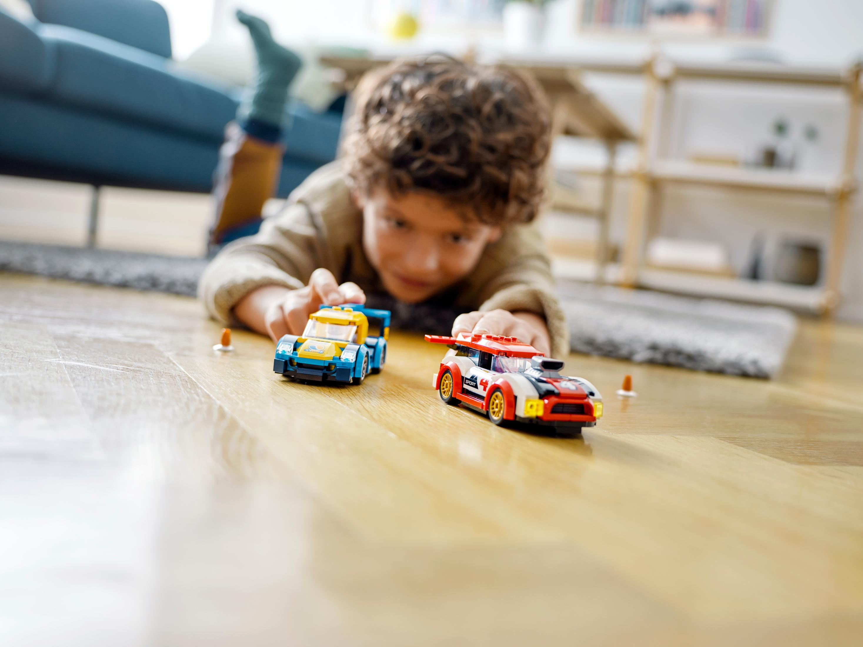 A child playing with two toy race cars on a wooden floor. One is blue, the other red.