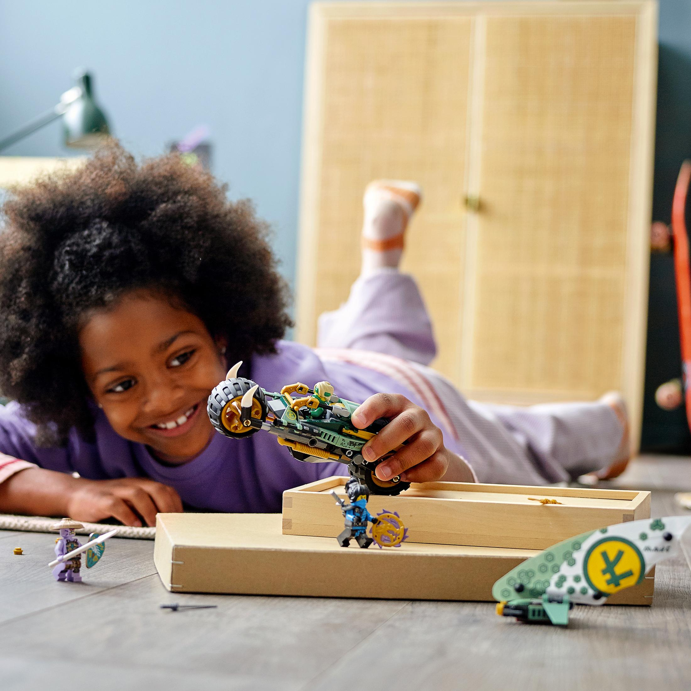 Ein Mädchen spielt mit Lego Ninjago Spielzeug. Es gibt Mini-Figuren und ein Fahrzeug. Der Hintergrund ist ein Raum.