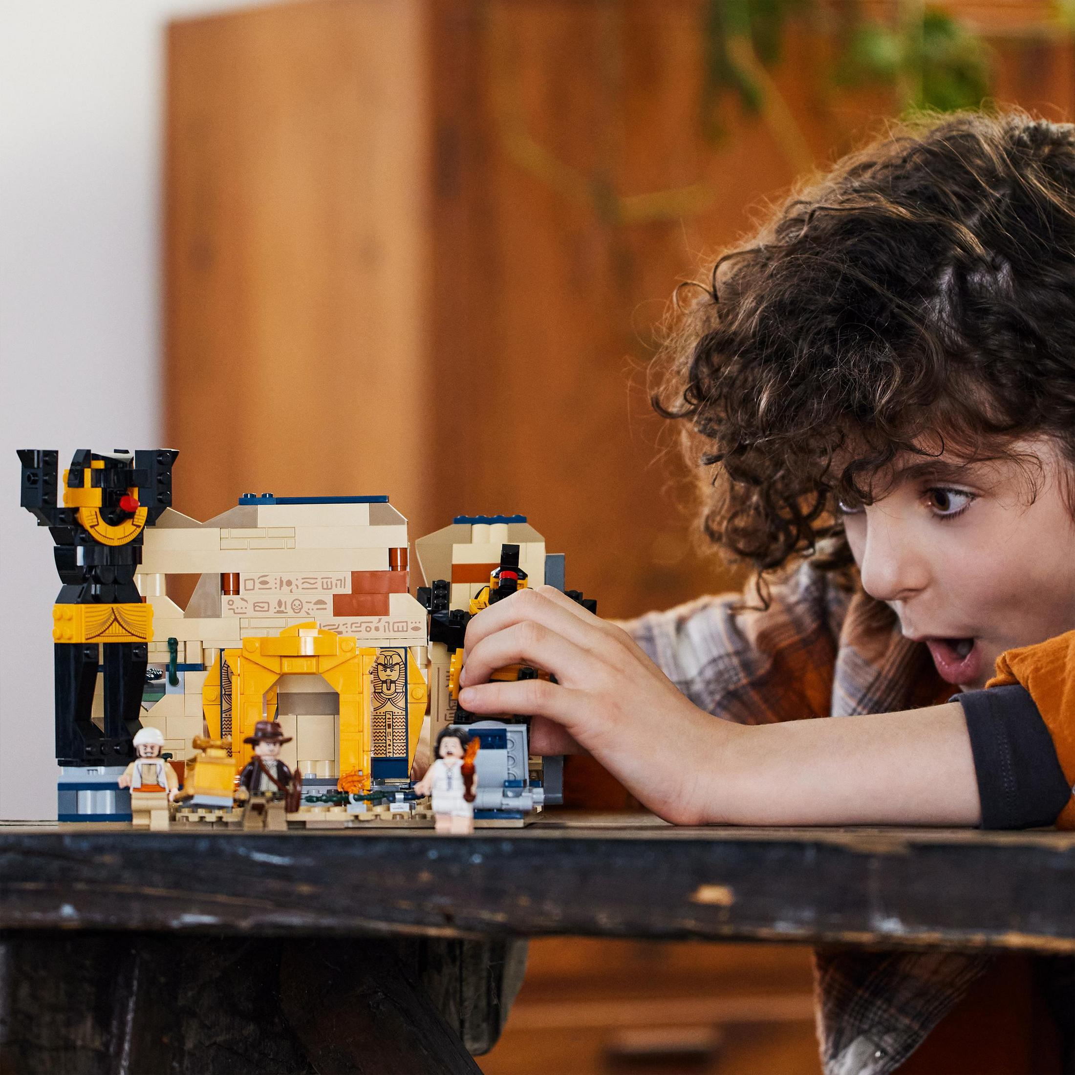 Un ragazzo guarda un set Lego di un antico tempio con personaggi, tavolo e sfondo in legno.
