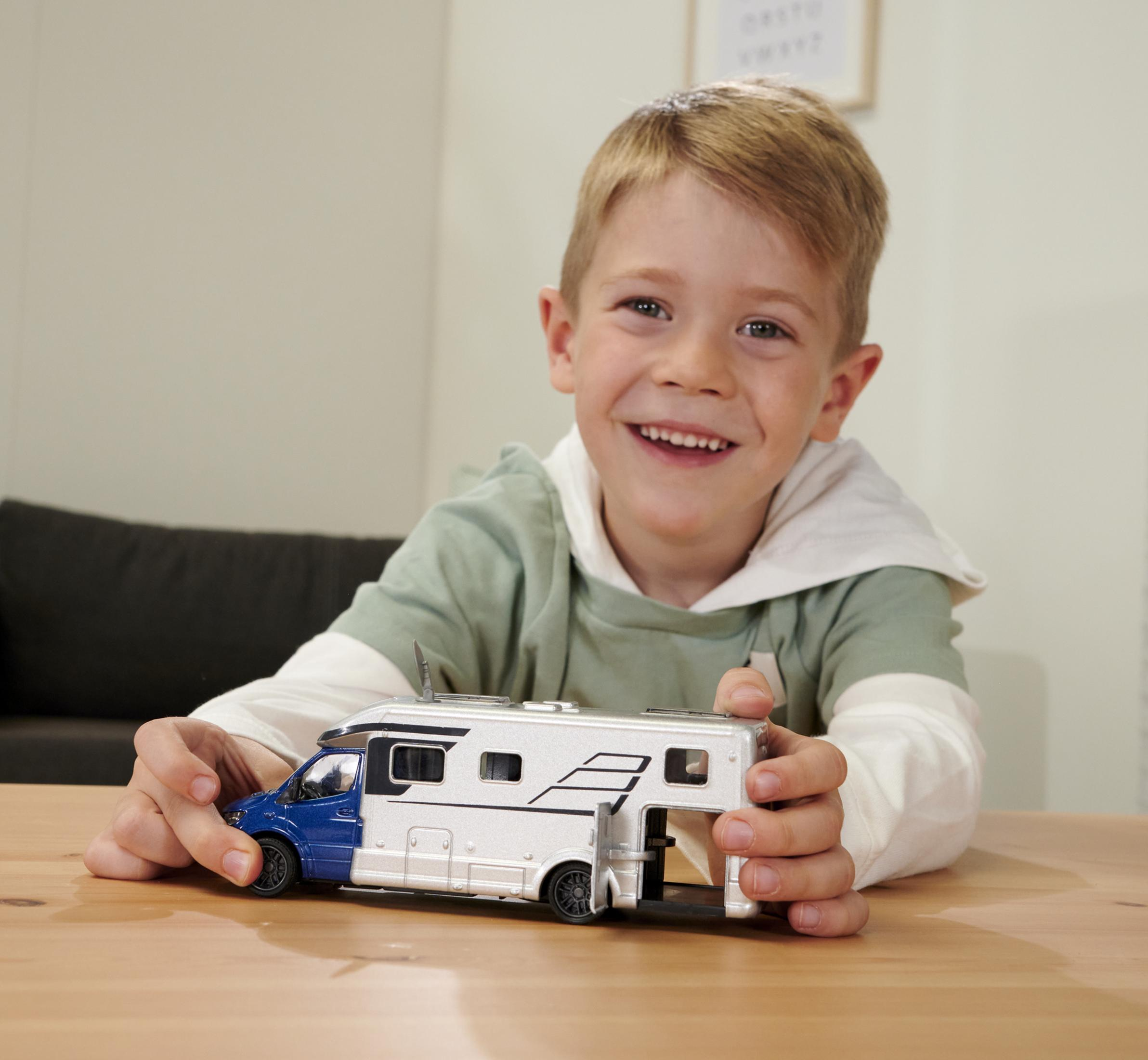 Mały chłopiec trzymający zabawkę kampera, siedzący przy drewnianym stole. Uśmiecha się.