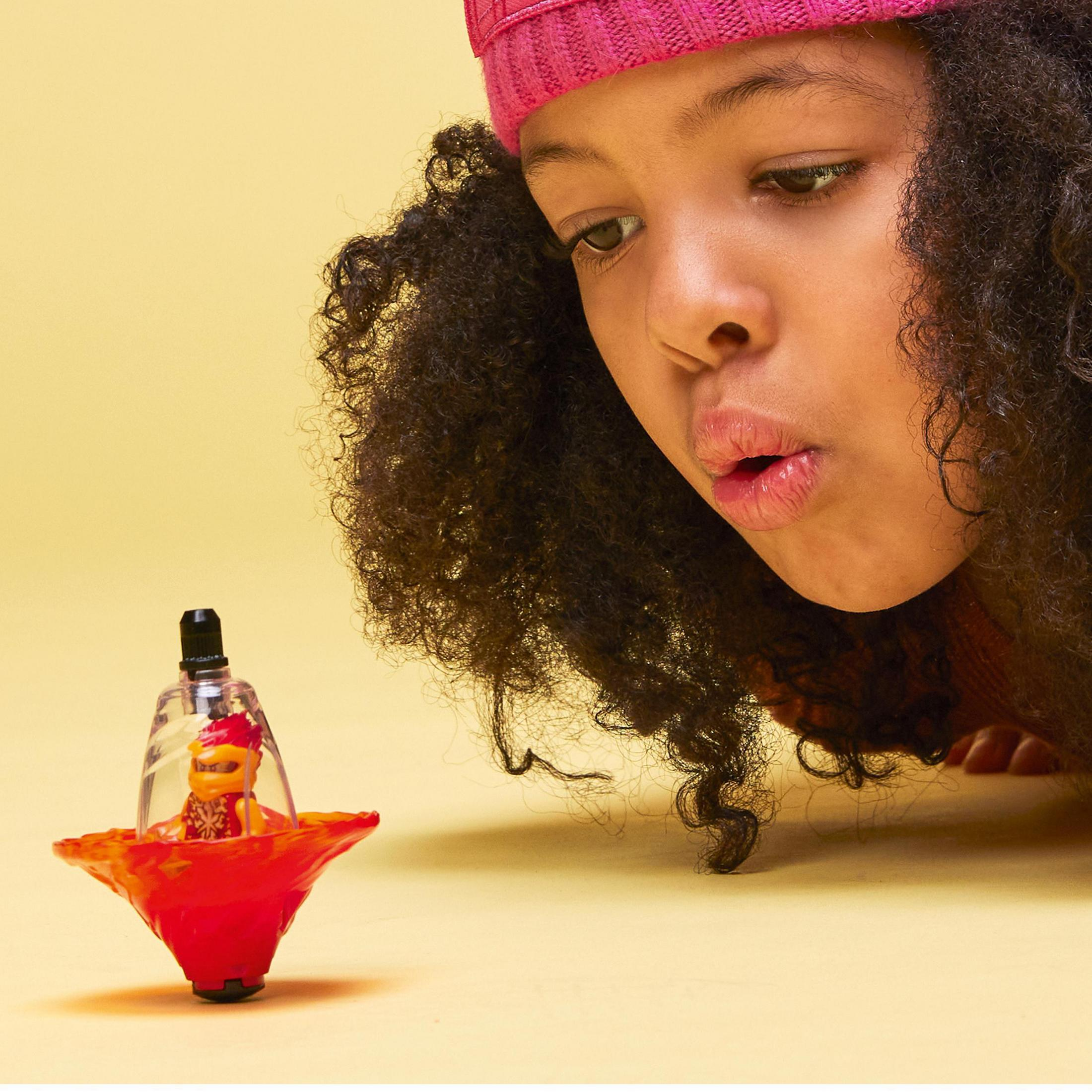 Une fille regarde un jouet toupie avec une petite figurine à l'intérieur, sur fond jaune.