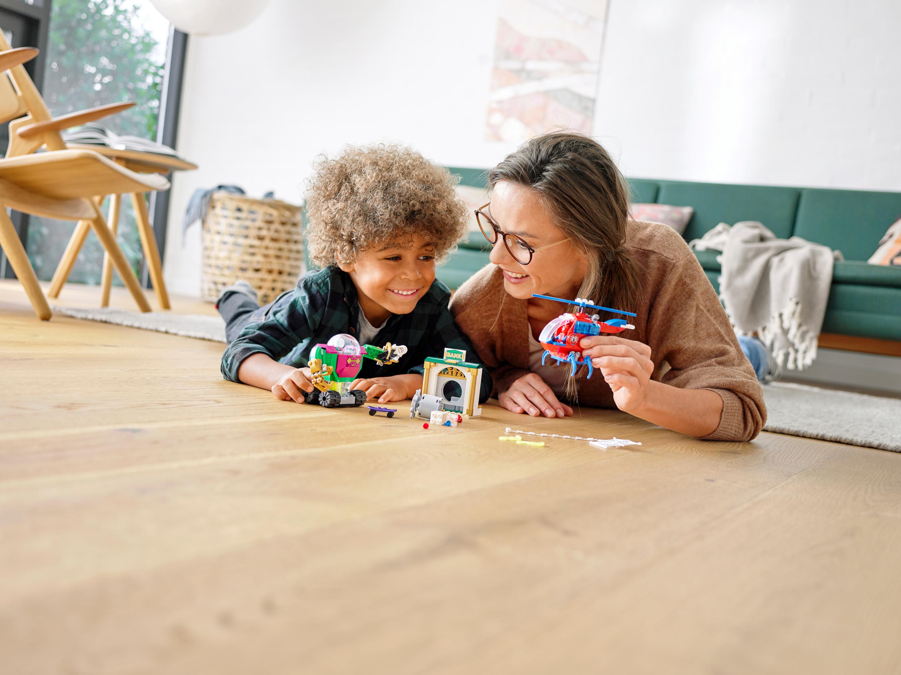 Ein Junge und eine Frau liegen auf einem Holzboden und spielen mit Lego-Spielzeug. Im Hintergrund ein grünes Sofa und ein Stuhl.