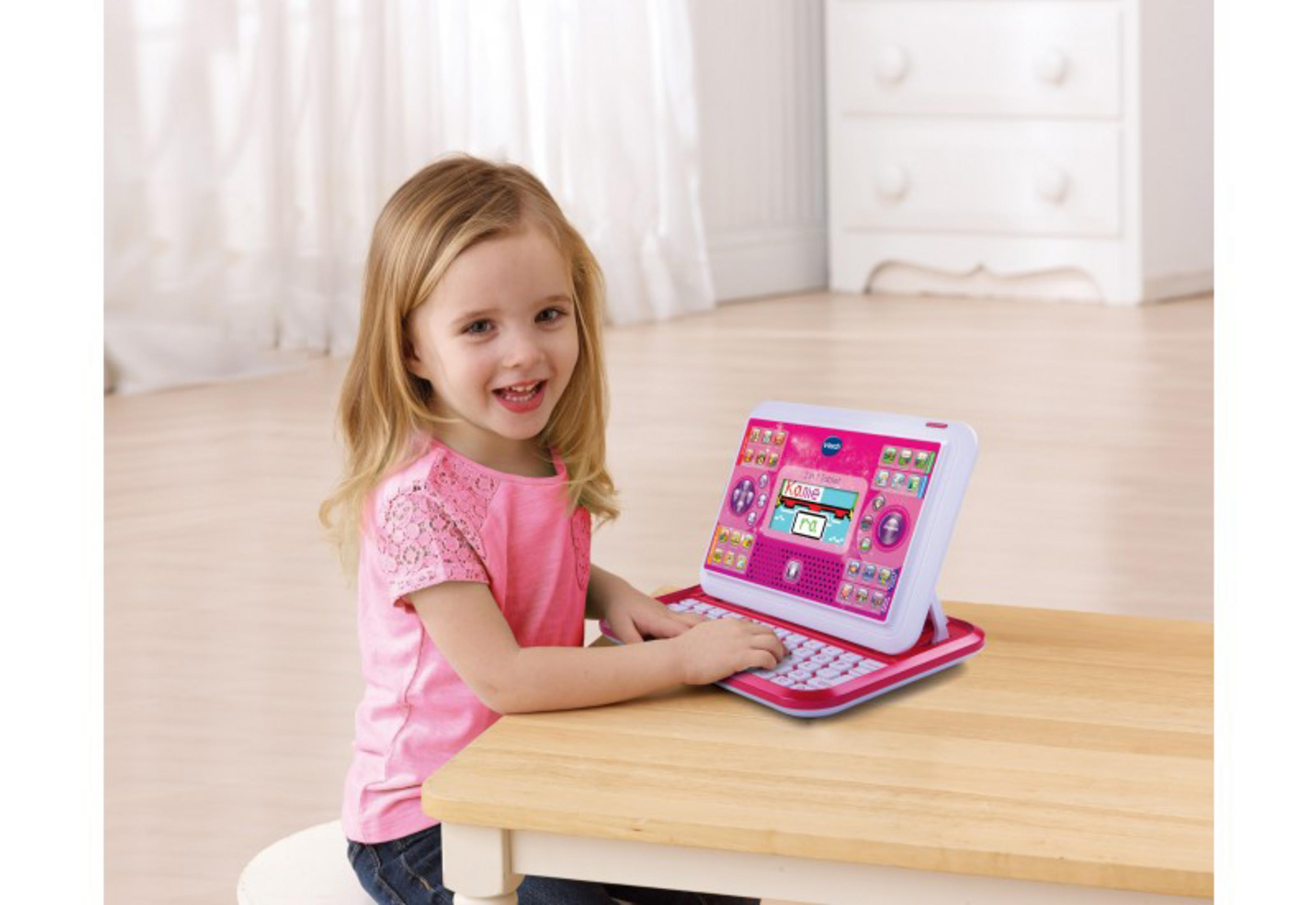 Ein junges Mädchen sitzt an einem Tisch und lächelt. Sie benutzt einen rosa und weißen Spielzeug-Laptop.