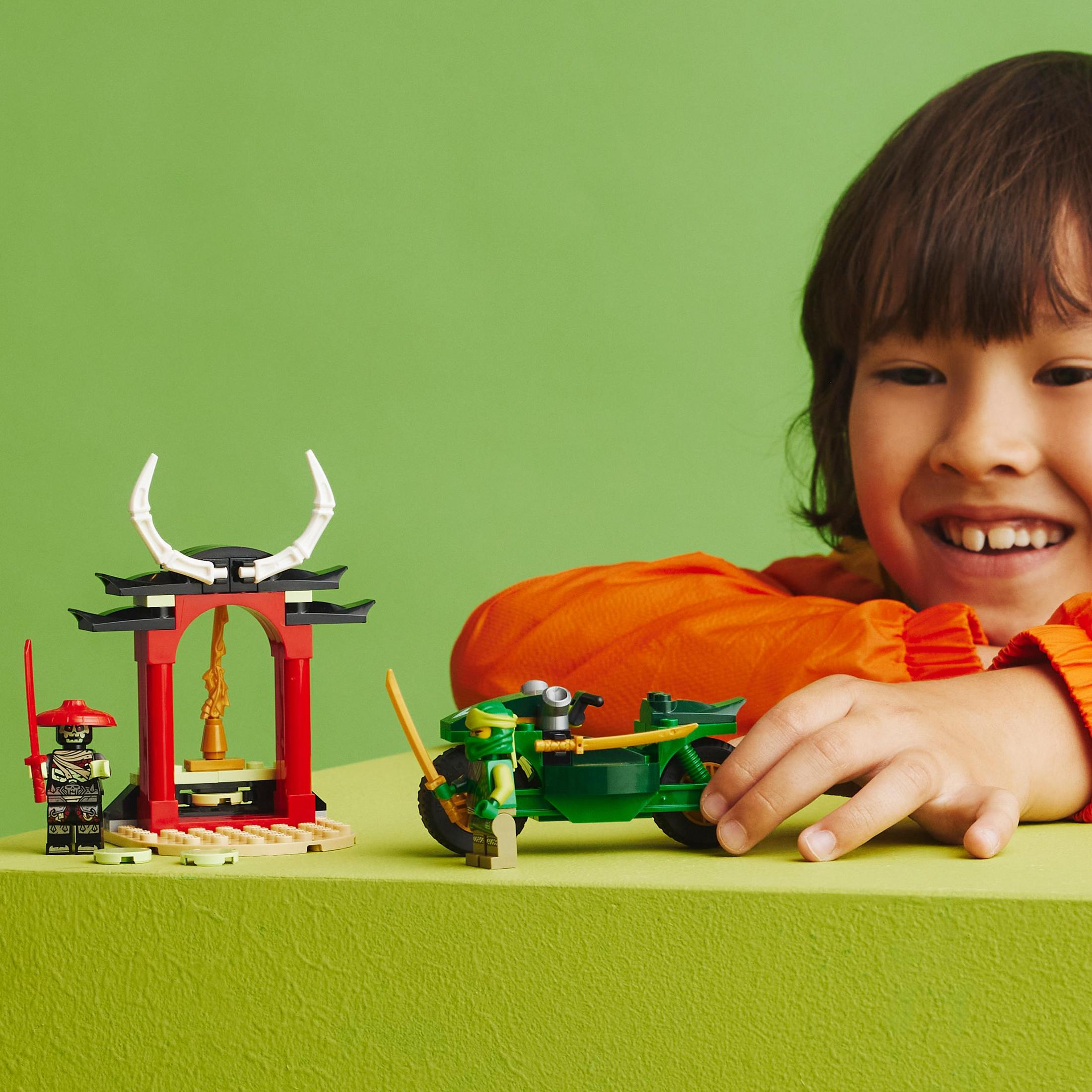 Bir çocuk gülümsüyor, yeşil bir arka plan üzerinde bir ninja ve bir motosiklet Lego seti ile.