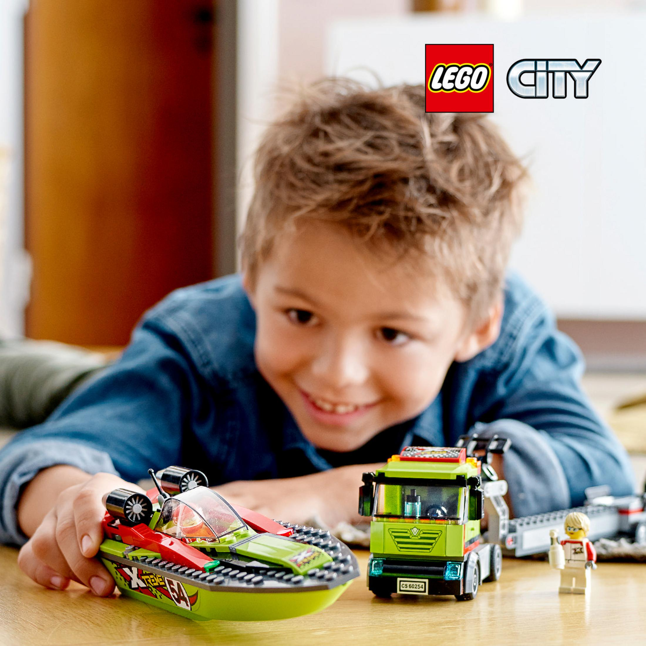 Un ragazzino sorride mentre gioca con una barca LEGO City verde e rossa e un camion giocattolo.