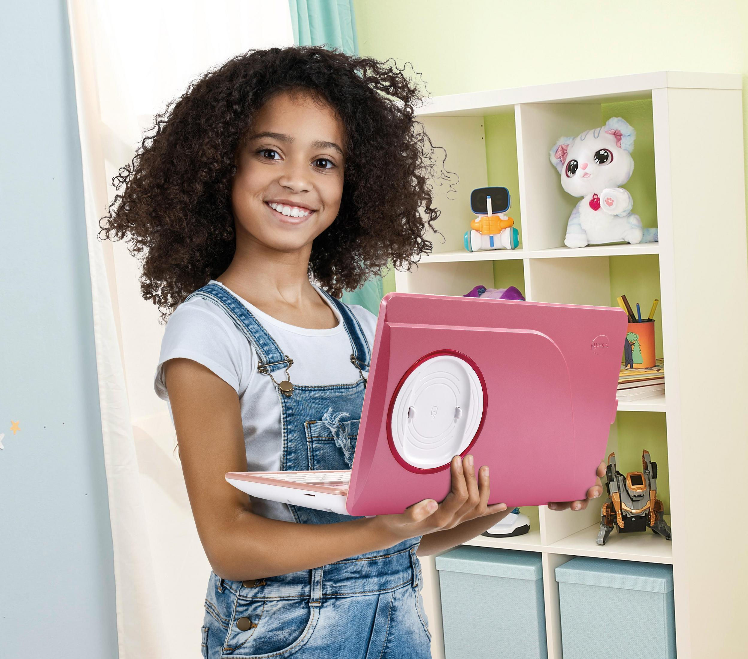 Mädchen mit lockigem Haar hält pinkfarbenen Laptop vor Bücherregal.