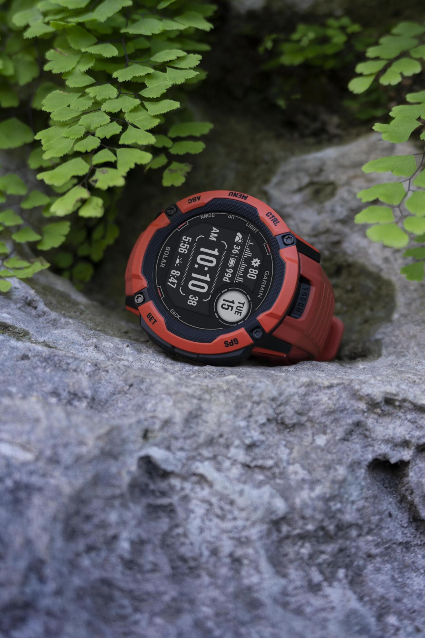 Een rode Garmin horloge ligt op een grijze rots. Groene varens op de achtergrond, onscherp.