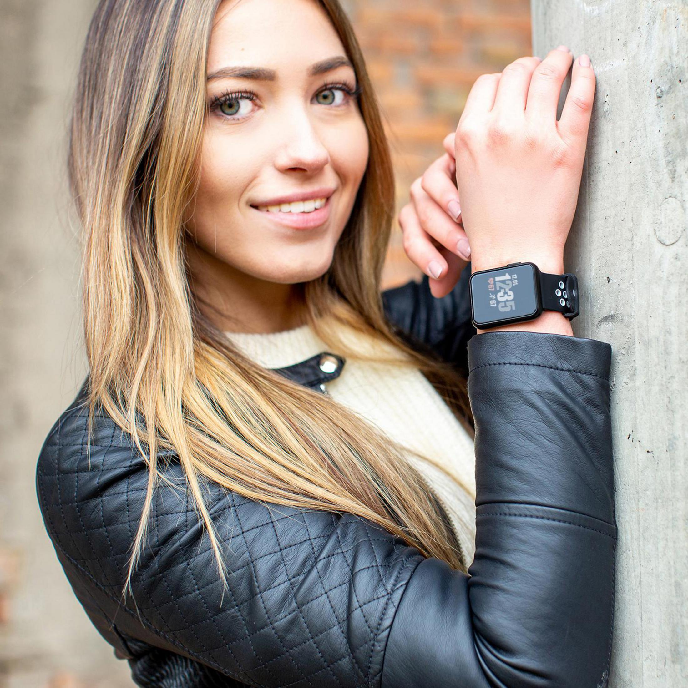 Eine Frau mit einer schwarzen Uhr am Handgelenk, lächelnd, lehnt an einer Betonmauer.