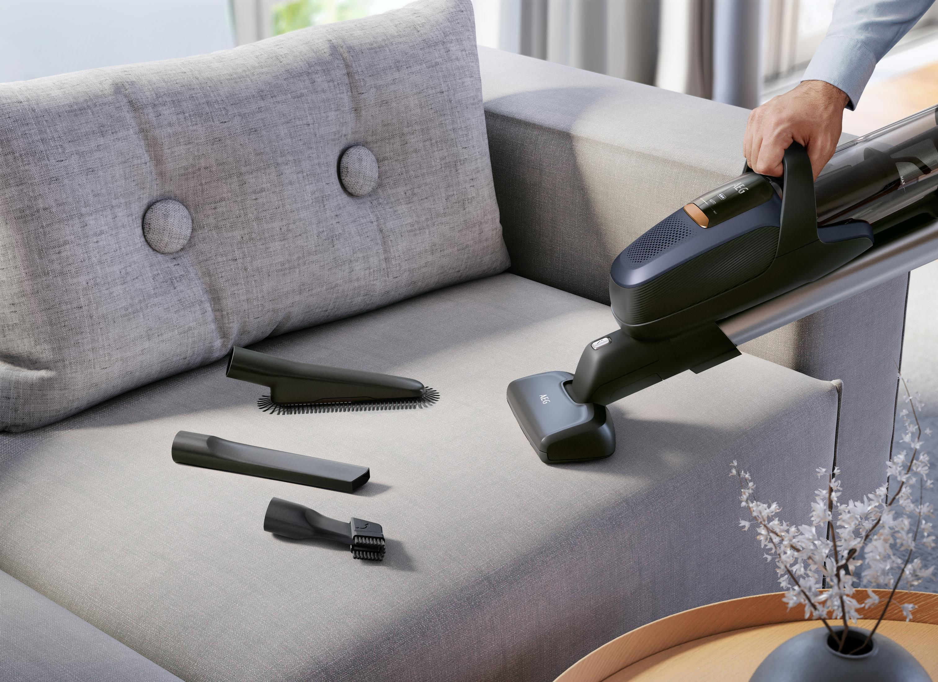 Osoba odkurzająca szarą sofę za pomocą nasadek. Istnieją dodatkowe nasadki.