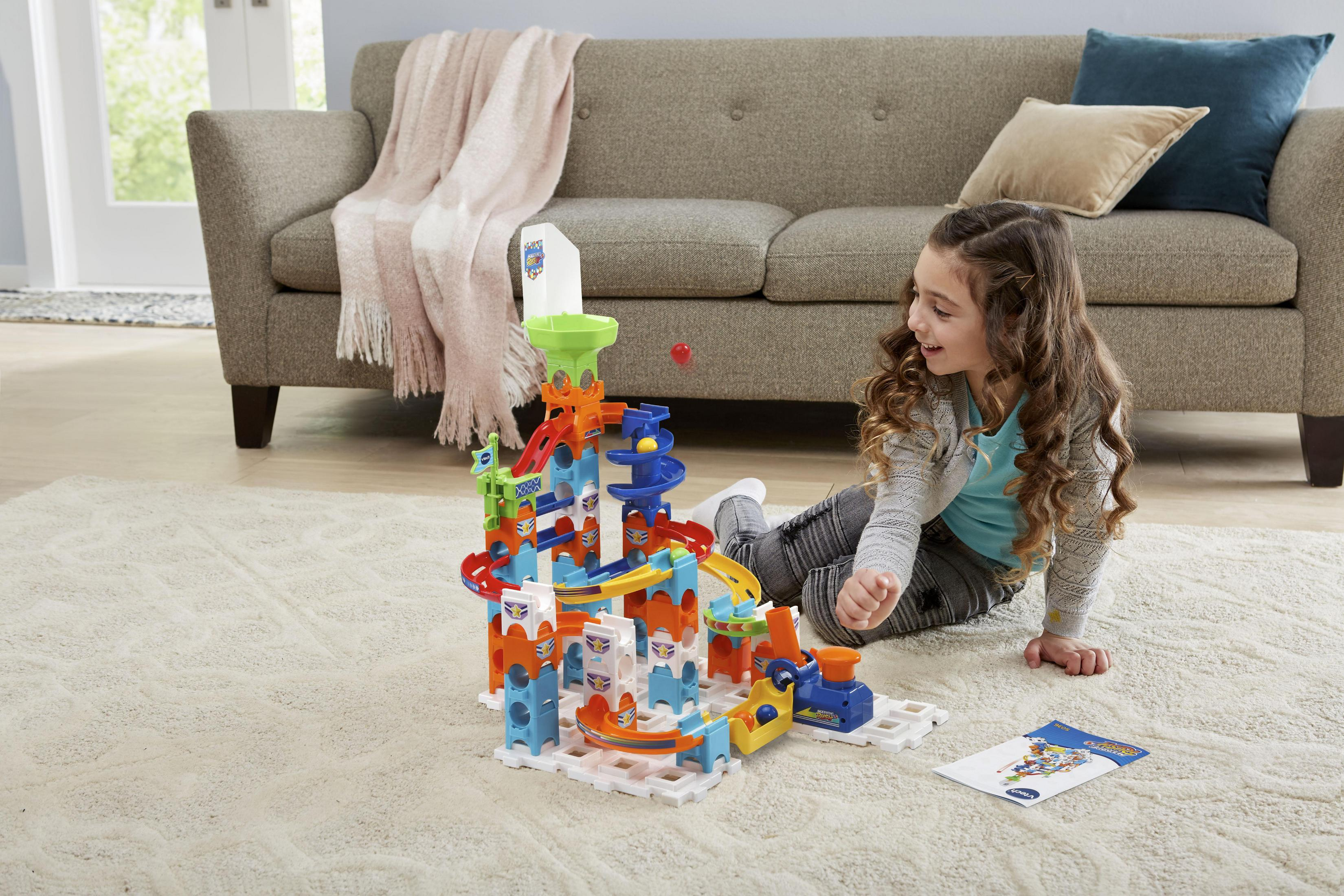 Mädchen spielt mit einem bunten Murmelbahnspielzeug auf einem gemusterten Teppich im Wohnzimmer.