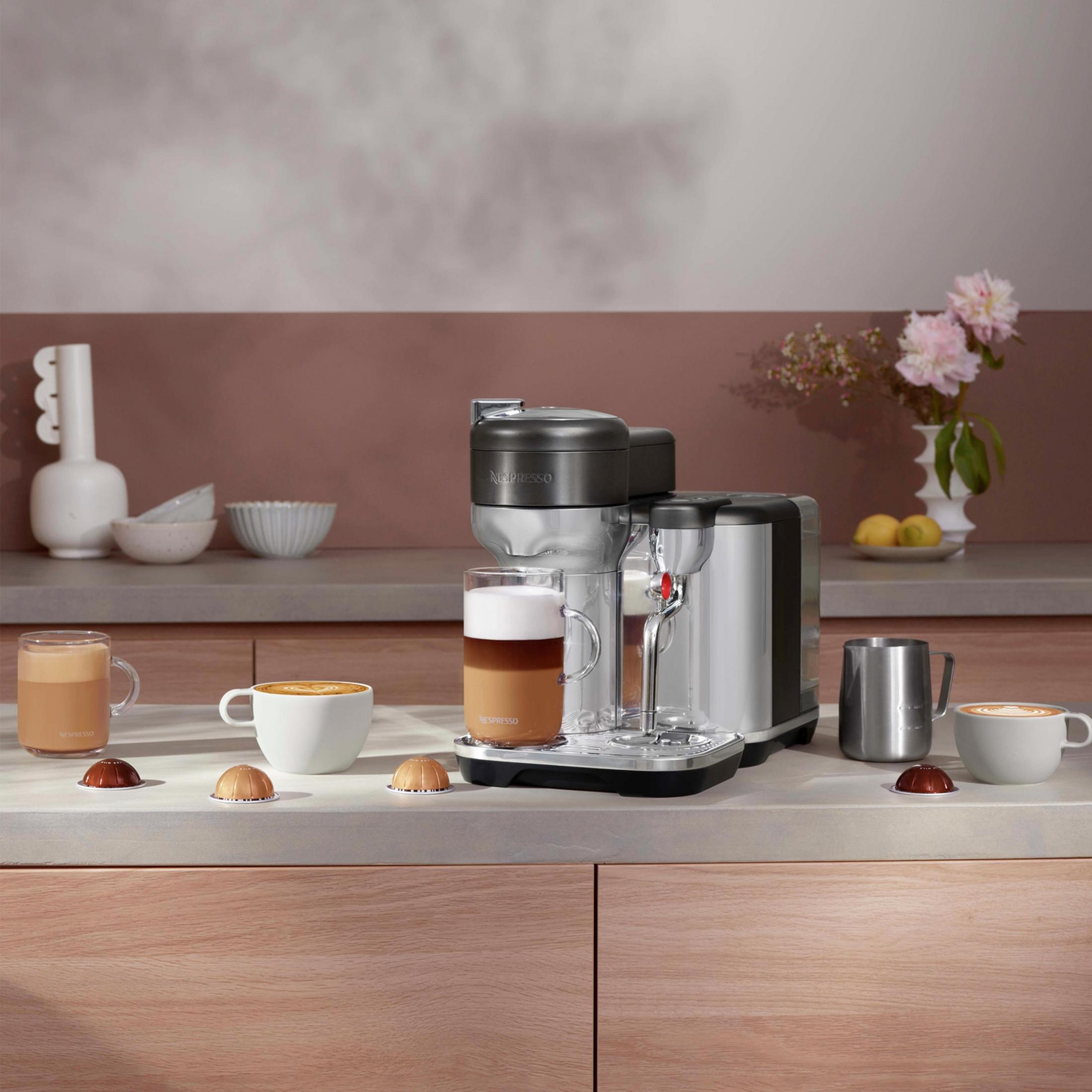 Eine Kaffeemaschine mit einem Glas Latte, Tassen und Kaffeepads auf einer Arbeitsplatte. Blumen sind im Hintergrund.