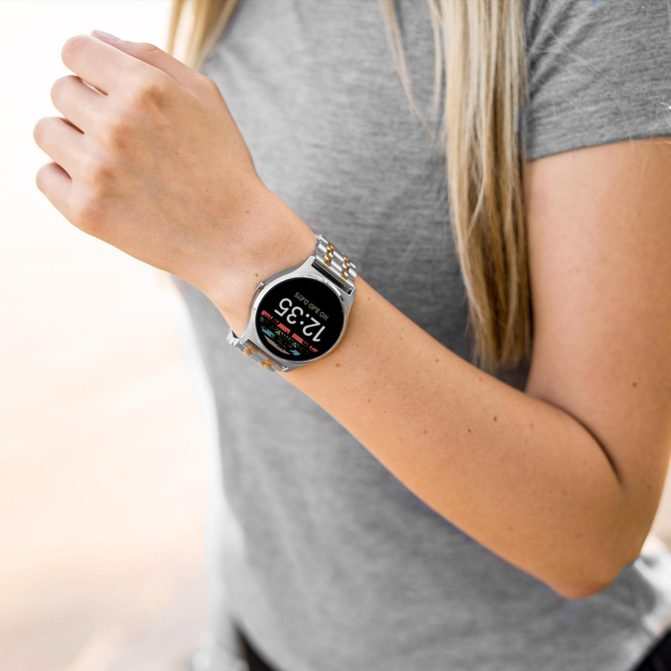 Ein Arm mit einer silber-goldenen Uhr, die 12:35 anzeigt. Sie trägt ein graues T-Shirt.