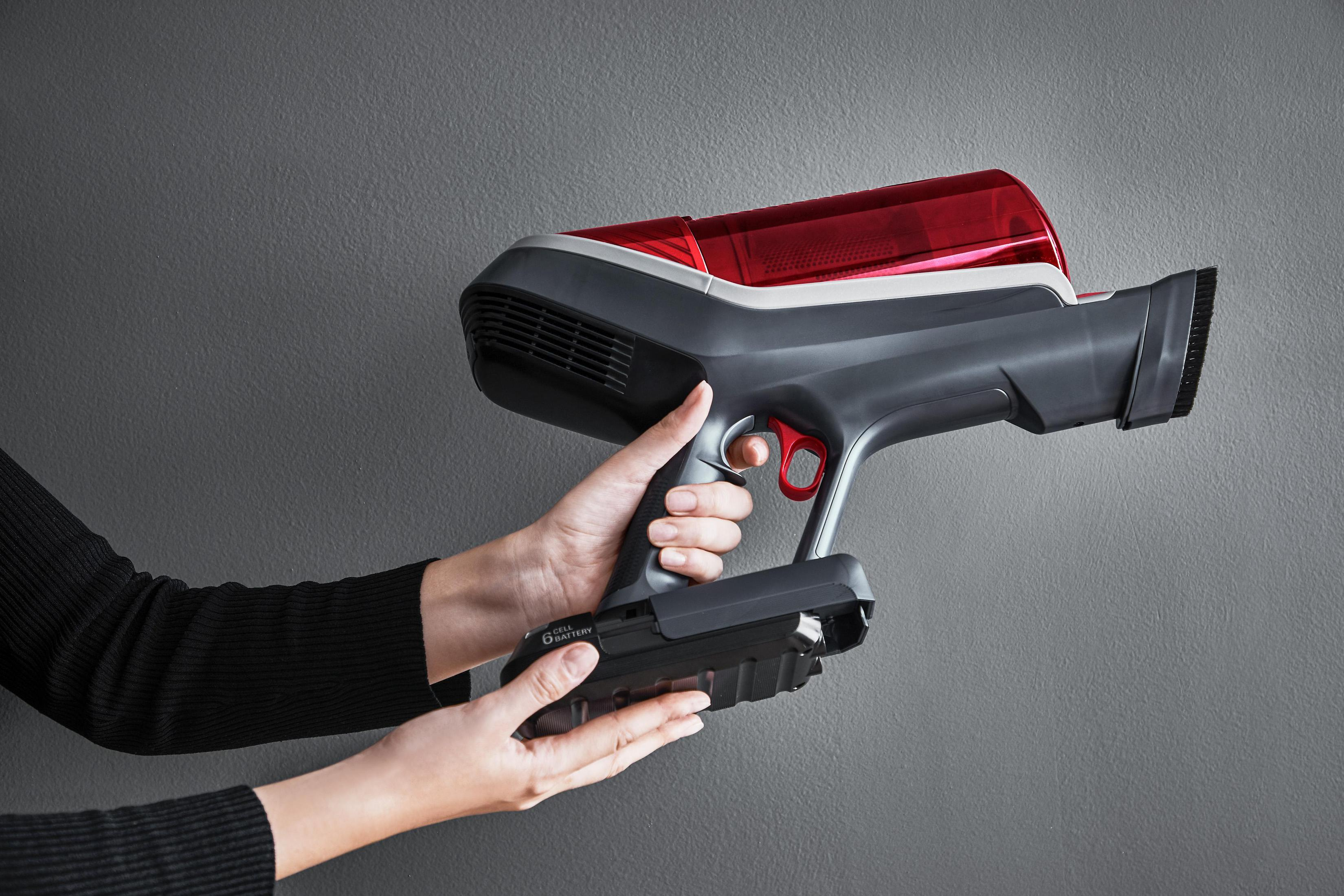 Osoba trzyma szaro-czerwony odkurzacz ręczny na tle szarej ściany.
