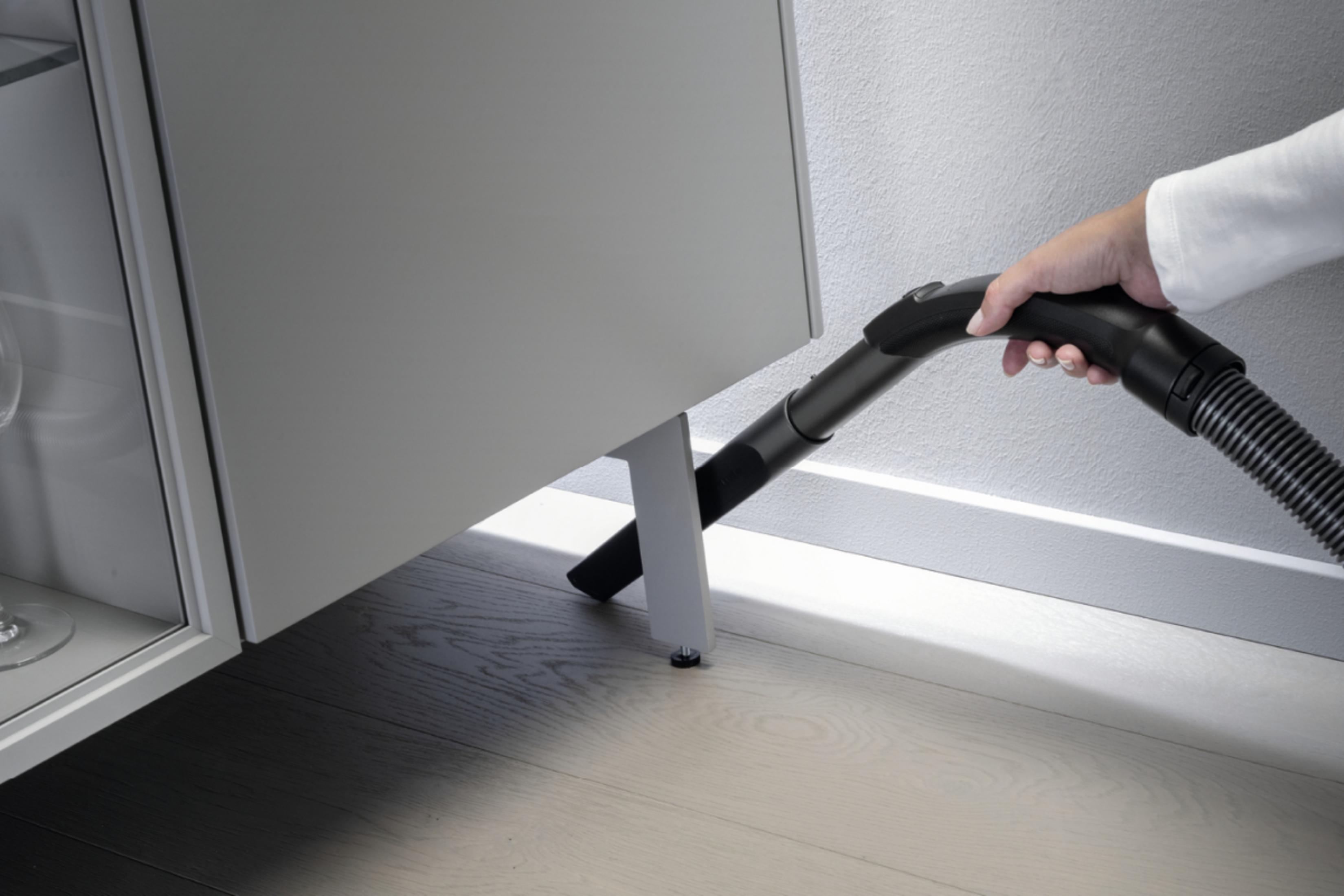 Eine Person, die mit einem schwarzen Staubsaugerschlauch unter einem grauen Schrank staubsaugt.